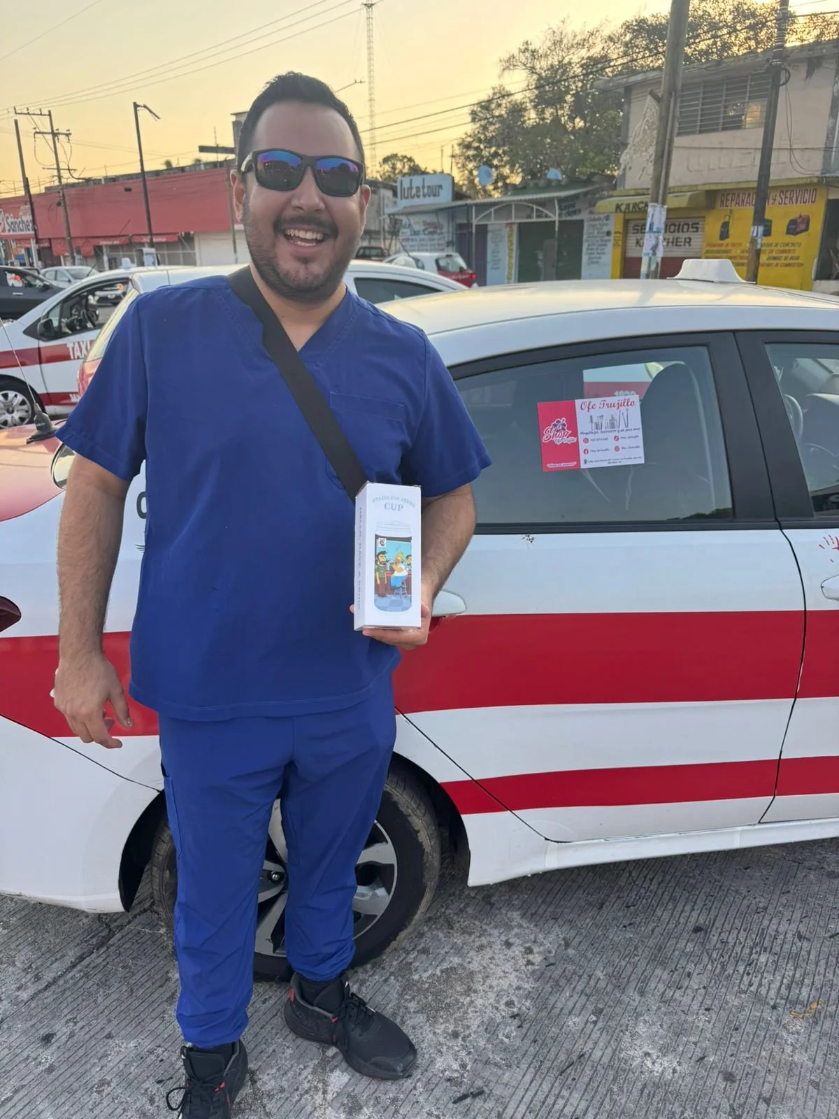 Hombre vestido con uniforme azul, con gafas de sol, sonriente, de pie frente a un coche de taxi, sosteniendo una caja de cerámica del Niño Jesús con la Virgen María en la parte superior. En el coche hay un cartel con información y la calle en un fond