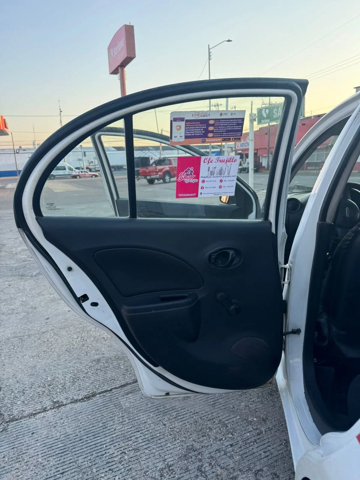Puerta abierta de un coche blanco con anuncios y stickers en la ventana trasera, en un estacionamiento con otros vehículos y señalización visible en el fondo.