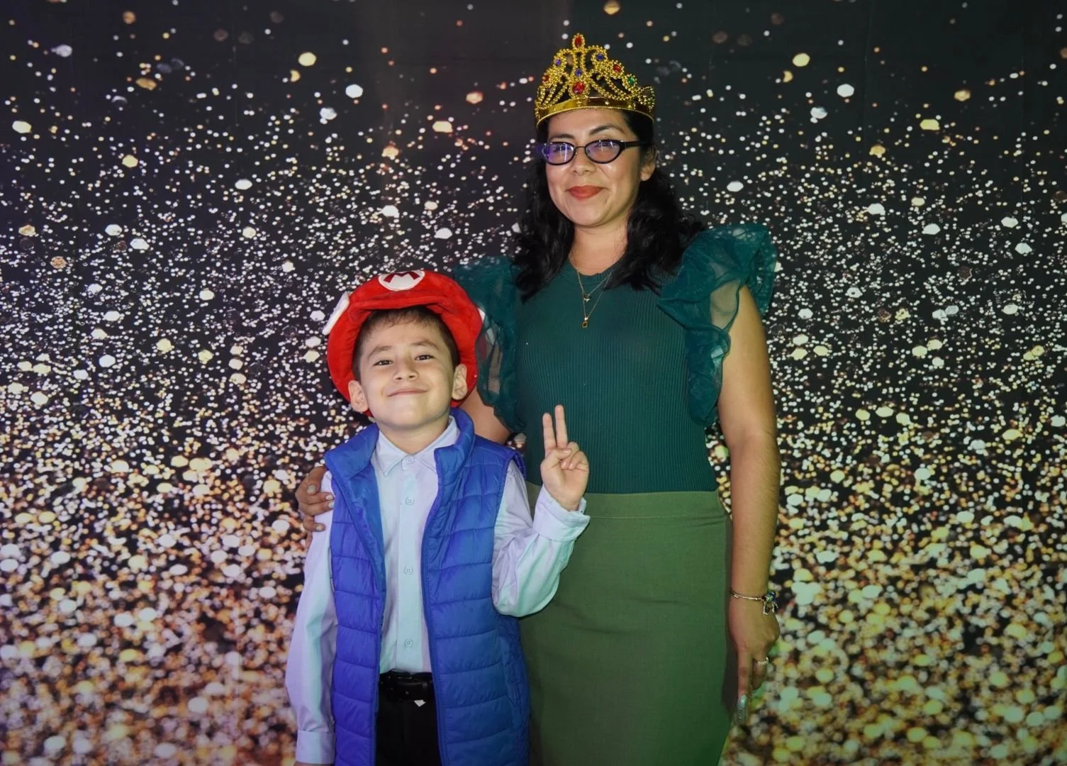 Una mujer con corona de fiesta y un niño con gorro de Mario Bros posando juntos frente a un fondo de luces doradas y blancas.