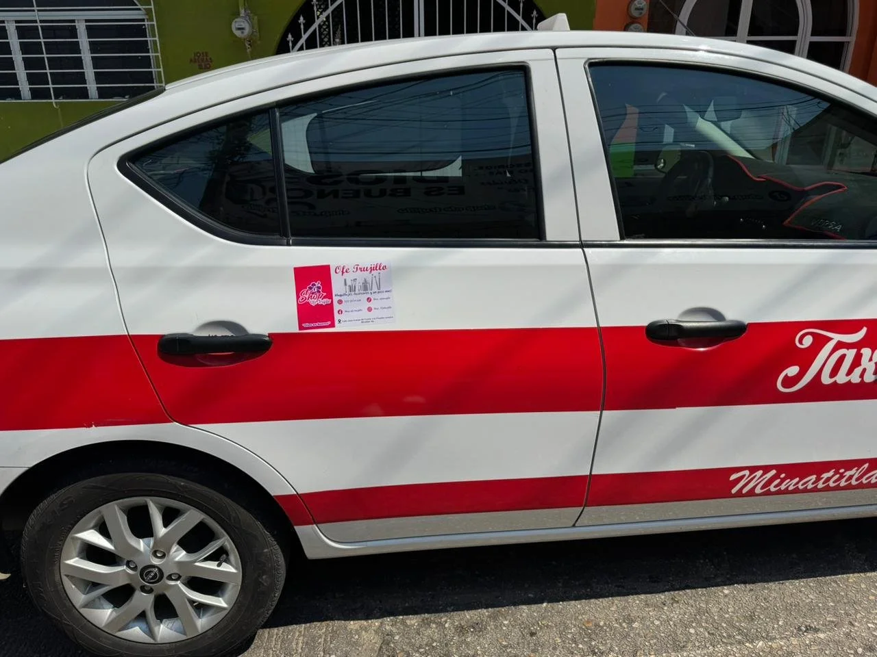 Auto de taxi con colores blanco y rojo, con un letrero pequeño en la ventana trasera izquierda que promociona un evento en Minatitlán.