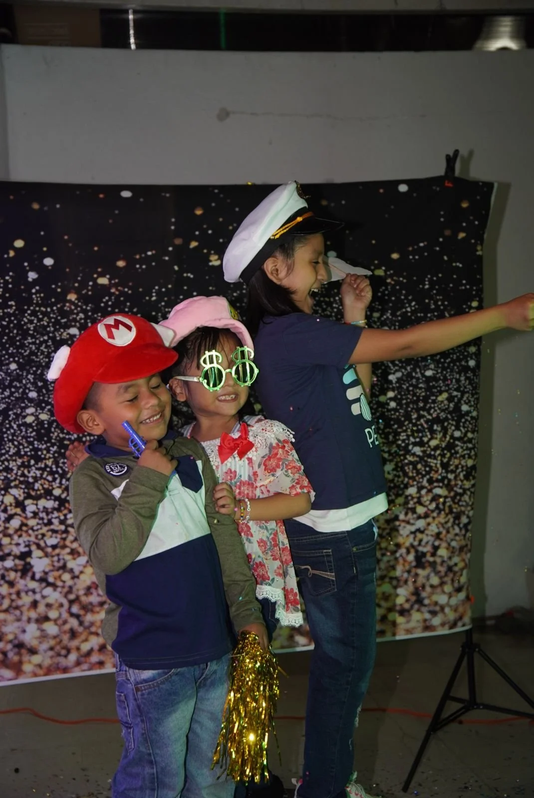 Tres niños con disfraces y accesorios de fiesta en un escenario con fondo de puntos blancos y negros.