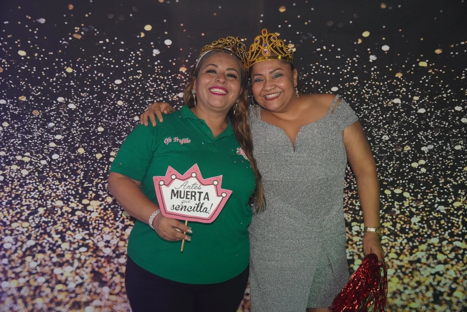 Dos mujeres sonrientes, una con corona y vestido plateado, la otra con camiseta verde y corona, en una celebración con fondo de confeti dorado.