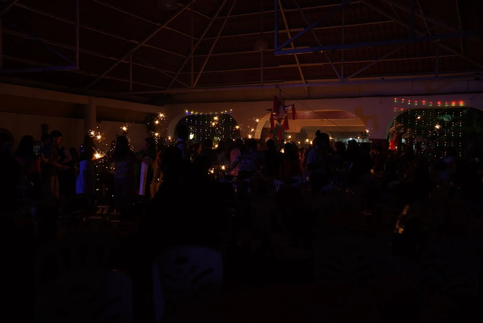 Celebración con muchas personas en un lugar cerrado, decorado con luces y adornos, con fuegos artificiales o fuegos artificiales de mano. En el centro hay una figura colgando, probablemente un elemento decorativo.