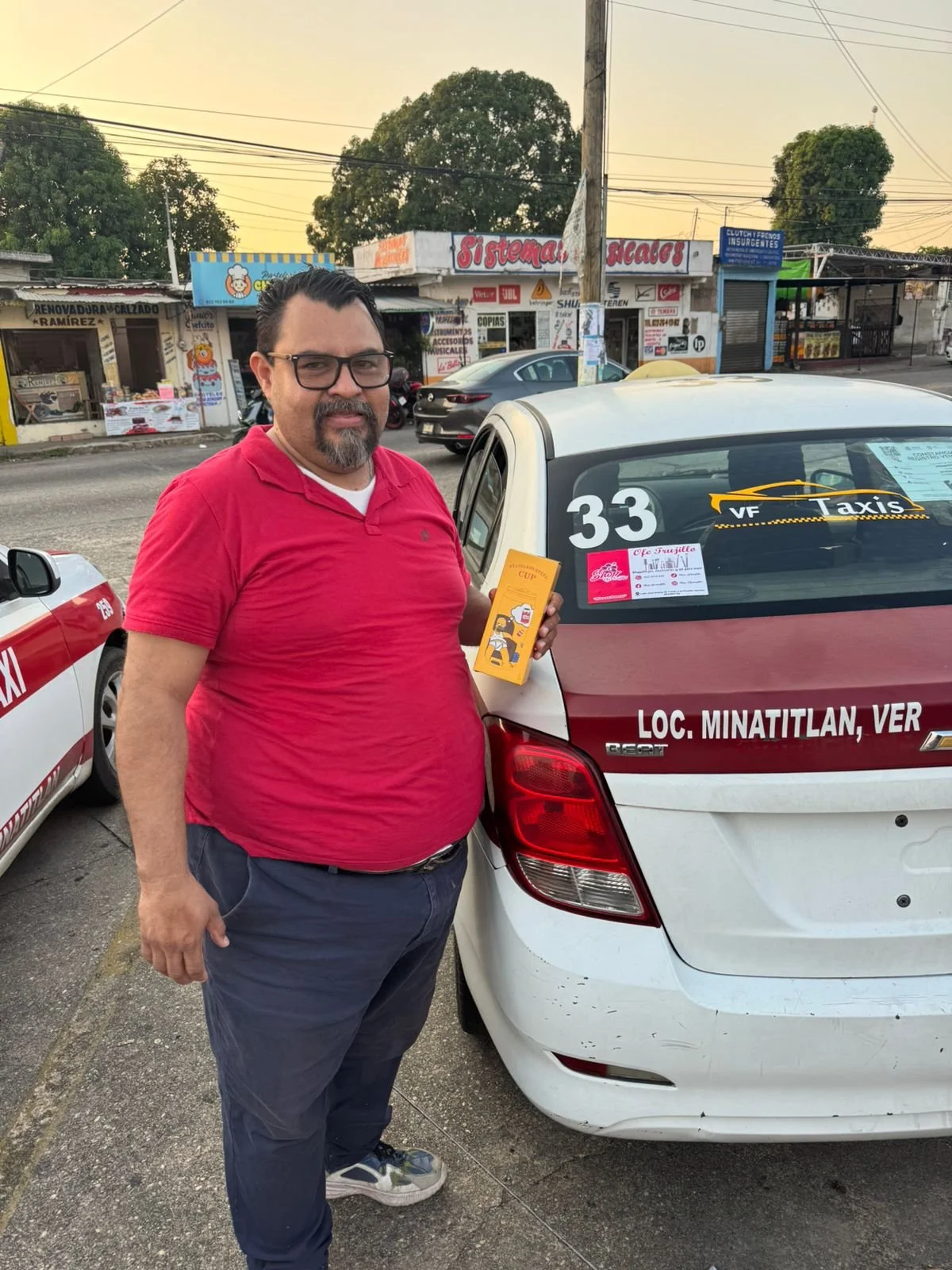 Hombre con gafas y barba, con camiseta roja, sosteniendo un paquete en la mano, de pie junto a un taxi en una calle con tiendas y árboles al fondo.