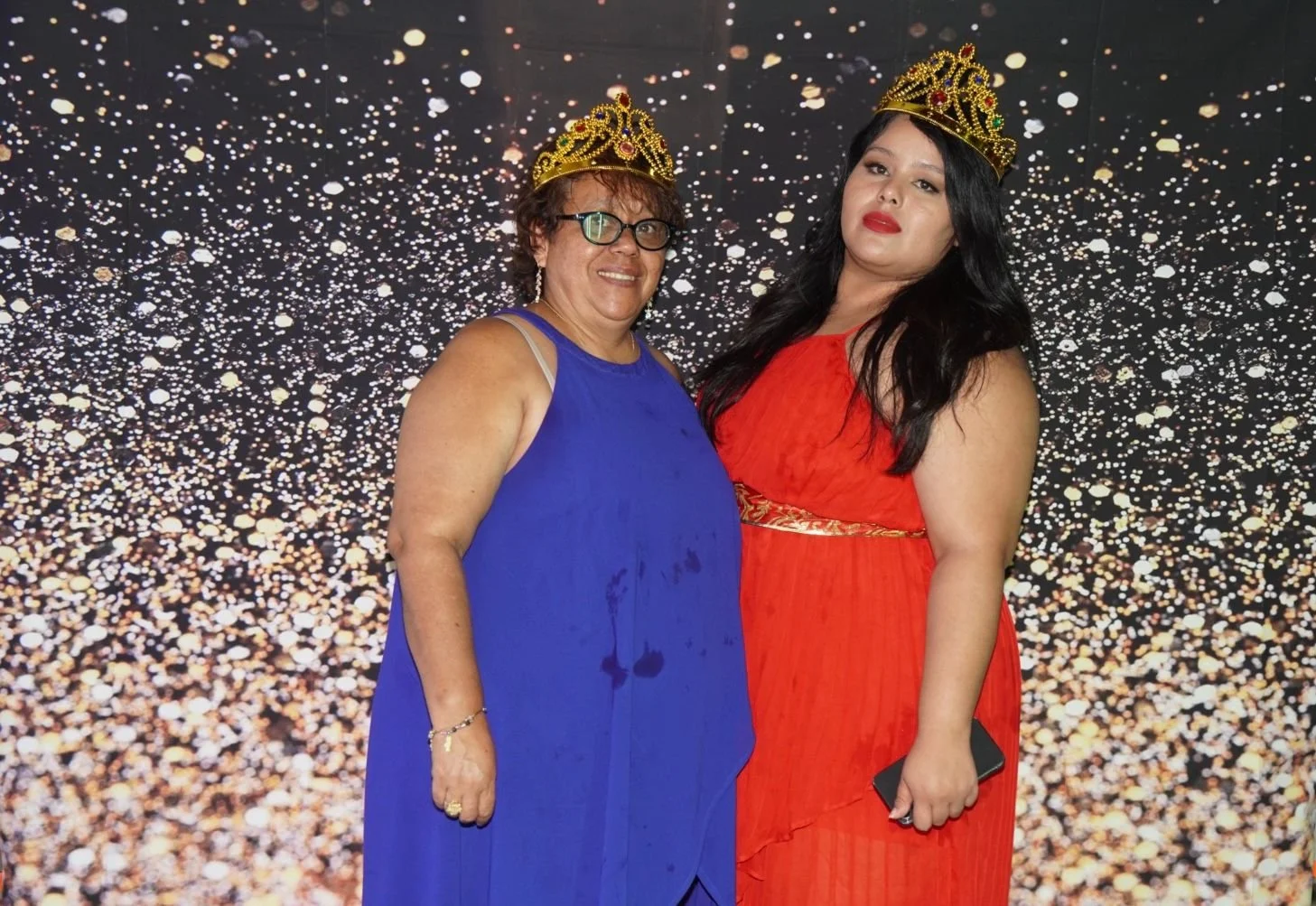 Dos mujeres usando coronas doradas, una con vestido azul y la otra con vestido rojo, posando frente a un fondo brillante de confeti dorado.