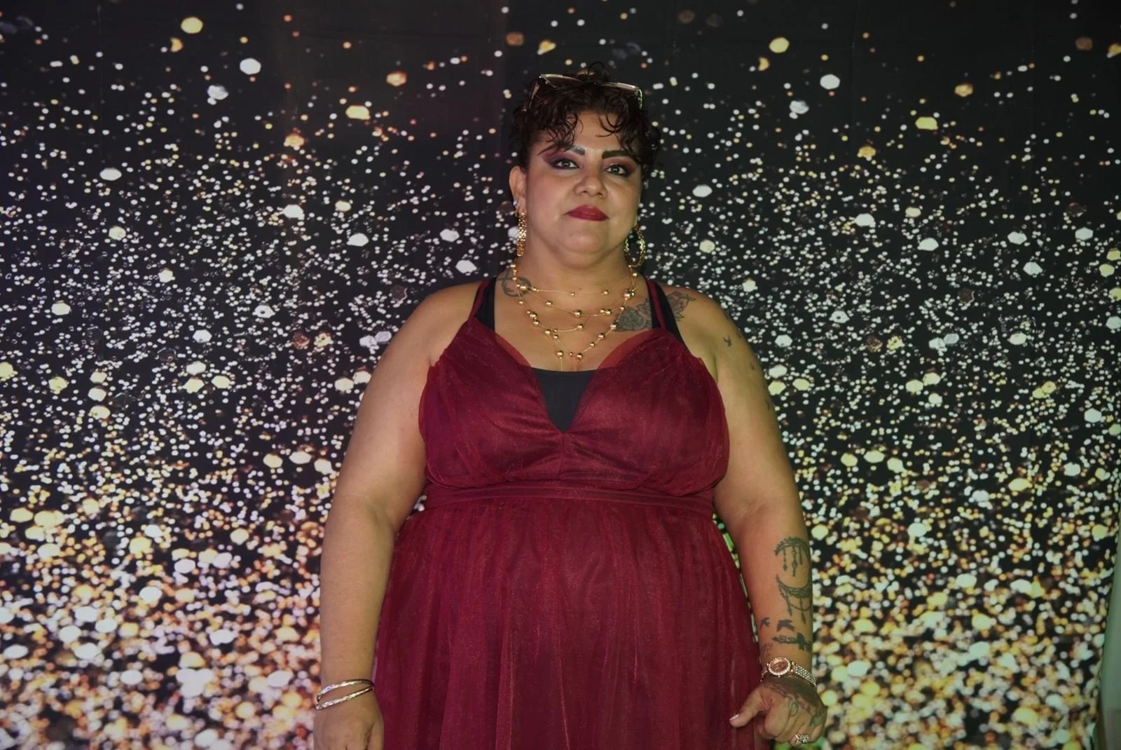 Mujer con vestido rojo y accesorios de oro posando frente a fondo de puntos dorados y blancos que simulan estrellas.