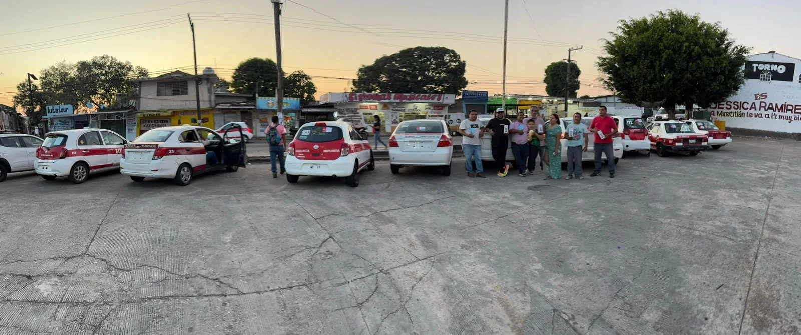 Grupo de personas posando frente a una fila de taxis blancos con franjas rojas en un estacionamiento en una ciudad, al atardecer.