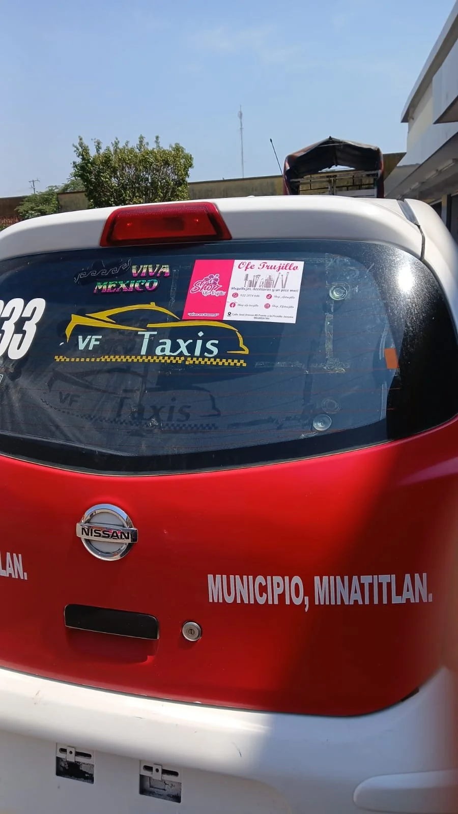 Parte trasera de un vehículo de taxi de color rojo y blanco con varios stickers, uno que dice VF Taxis, otro que dice Viva México y una publicidad de una tienda de cigarrillos llamada Ofe Trujillo.