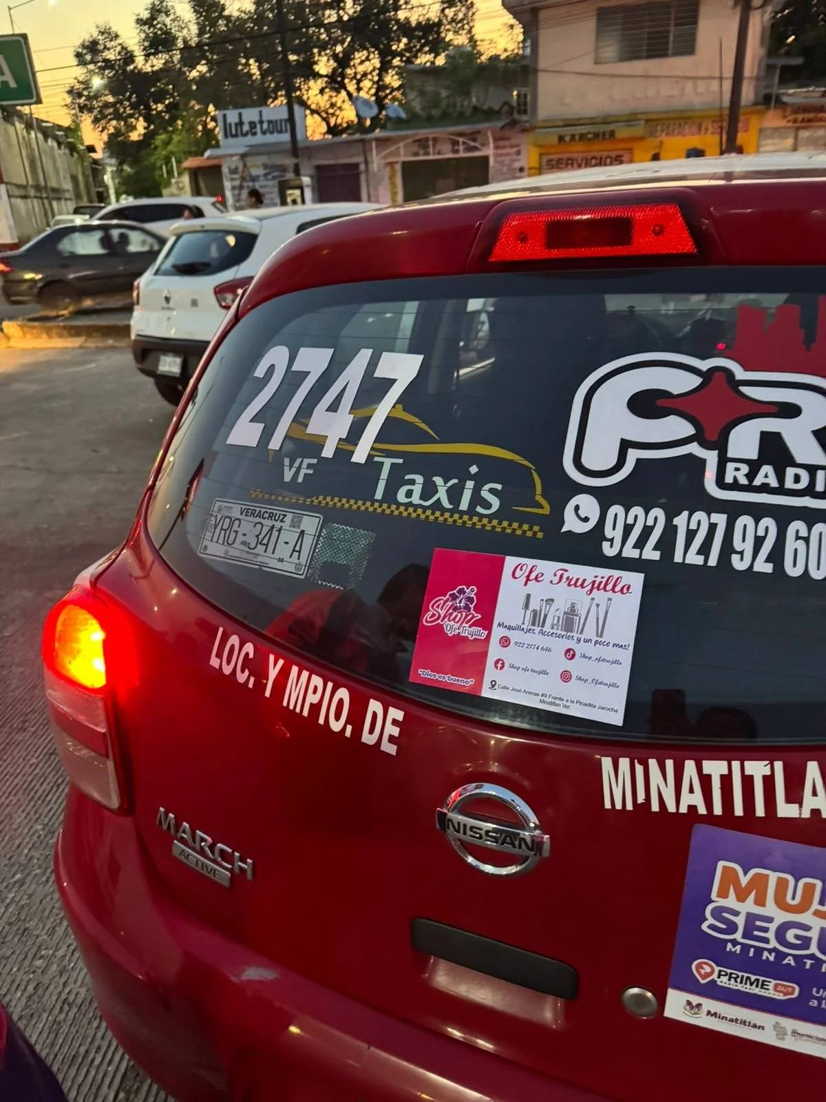 Imagen de la parte trasera de un coche Nissan March rojo con varias calcomanías y anuncios, incluyendo un letrero de Taxi con el número 2747 y publicidad para negocios locales en Veracruz.