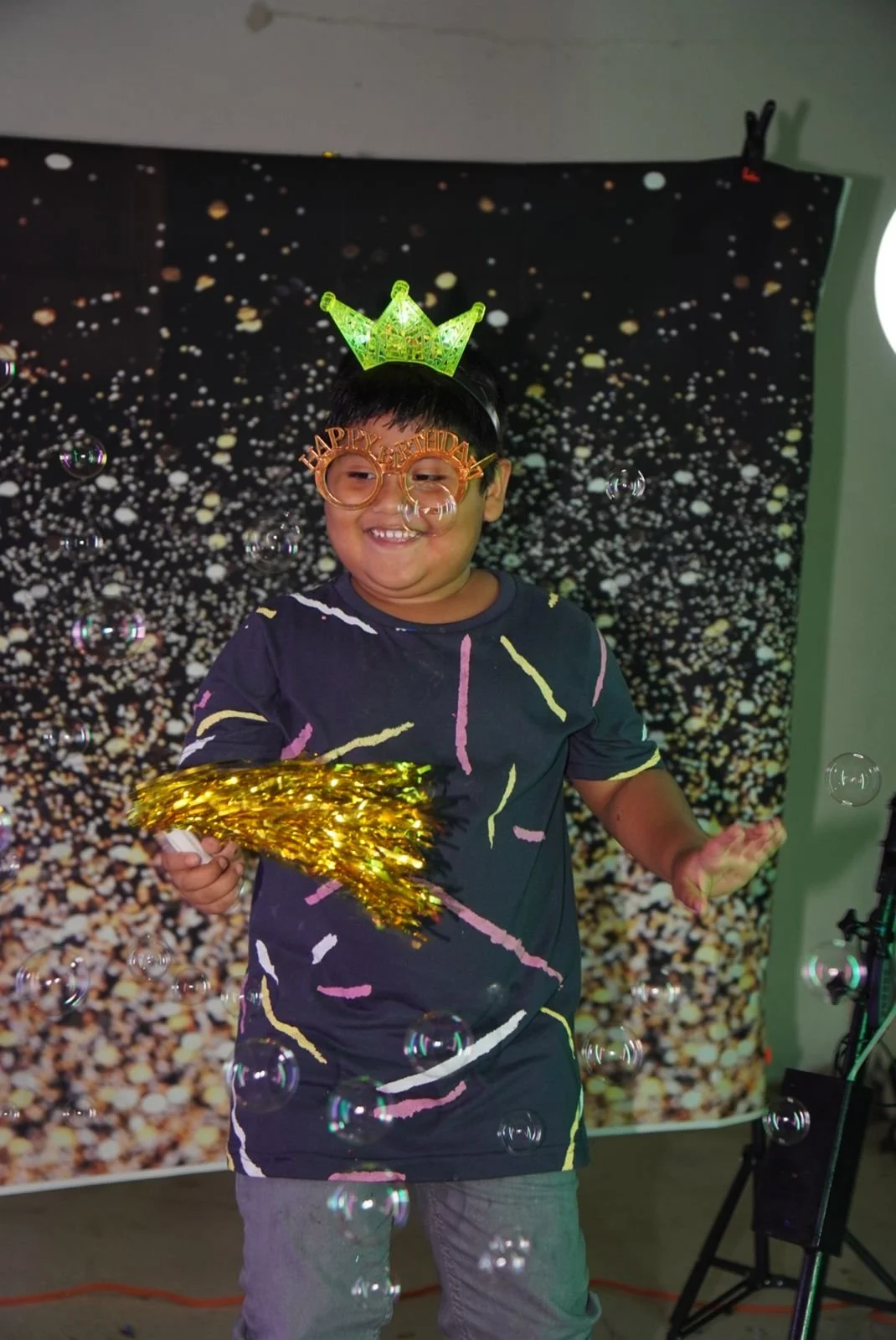 Niño en celebración de cumpleaños con gorro y gafas de fiesta, sosteniendo una corona de oro, rodeado de burbujas y un fondo decorativo de confeti.