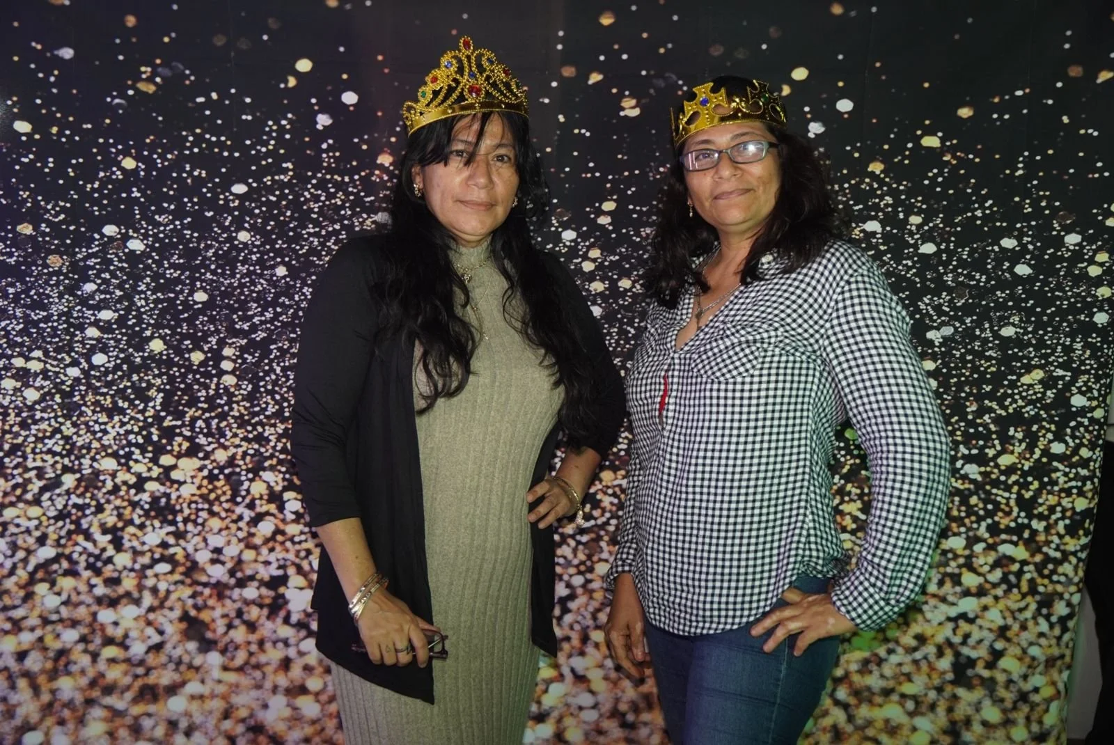 Dos mujeres con coronas doradas posando frente a un fondo con confeti dorado. La mujer a la izquierda lleva un vestido beige y un abrigo negro, y la mujer a la derecha lleva camisa a cuadros y gafas.