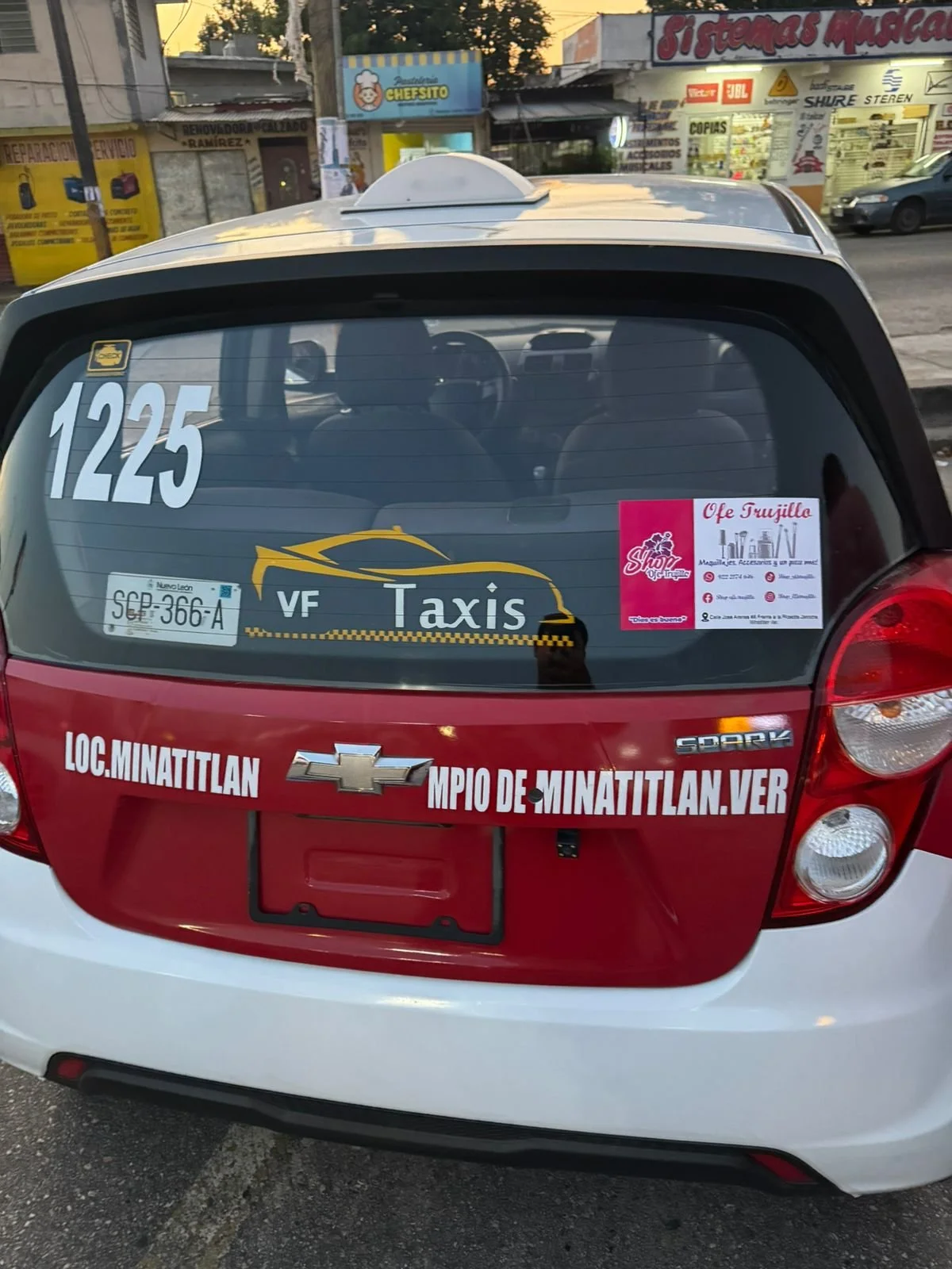 Parte trasera de un taxi Chevrolet Spark con logotipos, número, y anuncios en español."1225" y "VF Taxis" en la ventana trasera, y el texto "LOC. MINATITLAN MPIO DE MINATITLAN VER" en la parte trasera del coche.