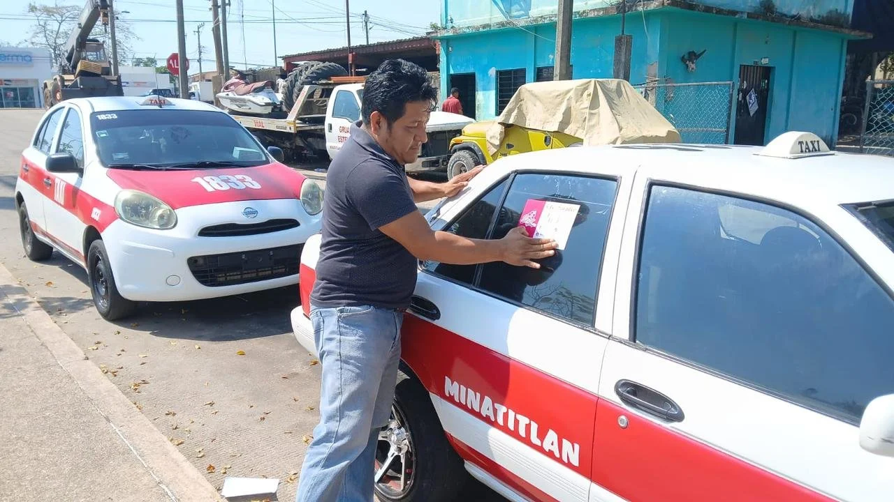 Un hombre colocando propaganda en un coche de taxi, que tiene letras que dicen 'MINATITLAN', en una calle. Al fondo hay otros vehículos y una casa azul.