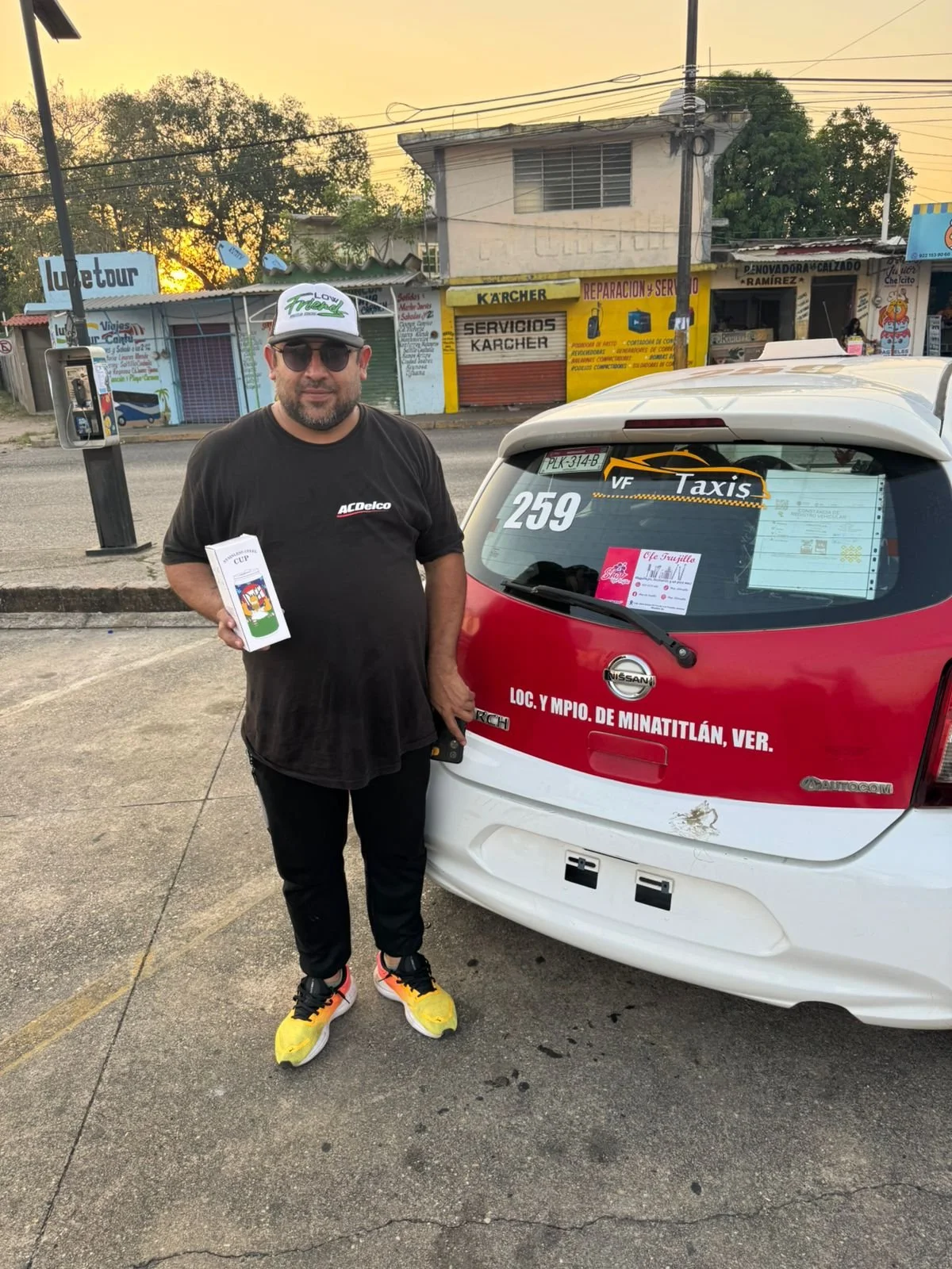 Hombre con gafas, sombrero y camiseta negra de pie junto a un taxi en una calle urbana al atardecer. El taxi es un Nissan con cartel que indica que pertenece a Minatitlán, Veracruz.