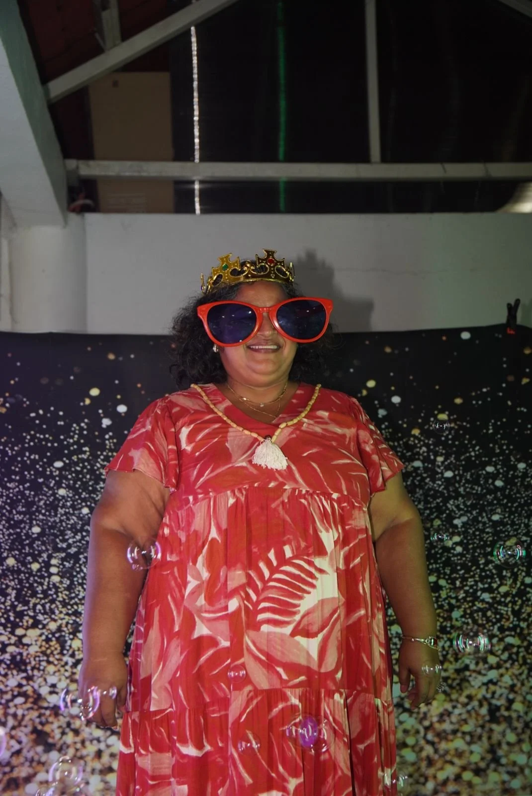 Mujer sonriendo con gafas grandes rojas, corona decorativa y vestido rojo con estampado de hojas, en un ambiente decorado con fondo de estrellas y burbujas.