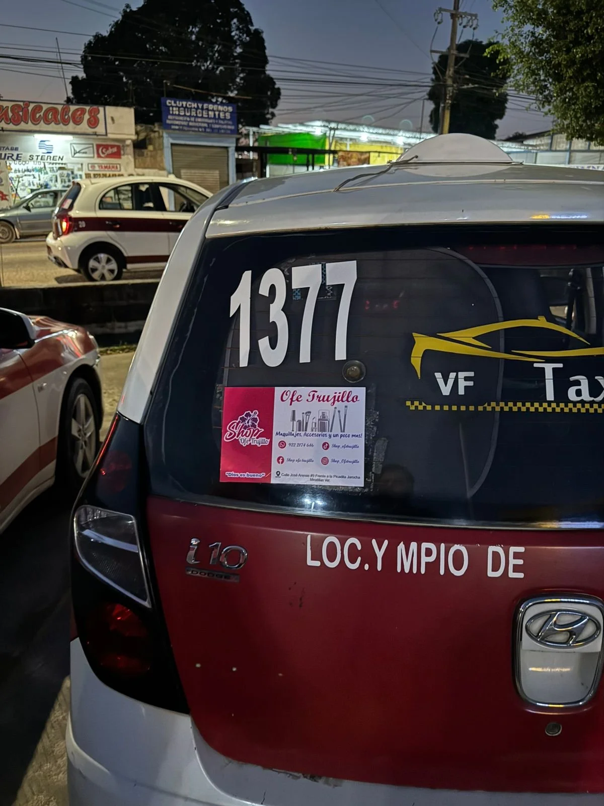 Parte trasera de un vehículo de taxi con el número 1377, en una calle con otros autos y anuncios en los locales comerciales.