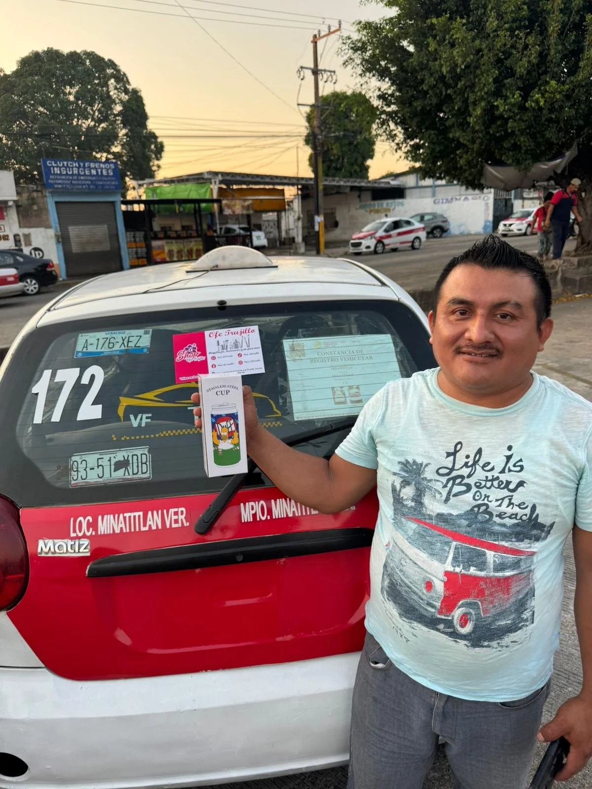 Un hombre sonriente sosteniendo una taza de cerámica frente a un automóvil con varios papeles y anuncios en la ventana trasera en una calle urbana durante el atardecer.
