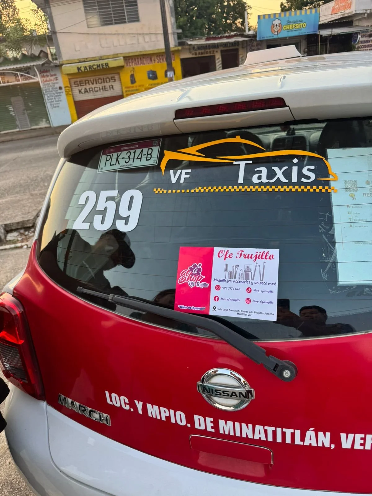 Parte trasera de un taxi Nissan con anuncios y señalización en la ventana y carrocería, incluyendo un cartel de una tienda llamada 'Shop Ofe Trujillo' y detalles de ubicación.