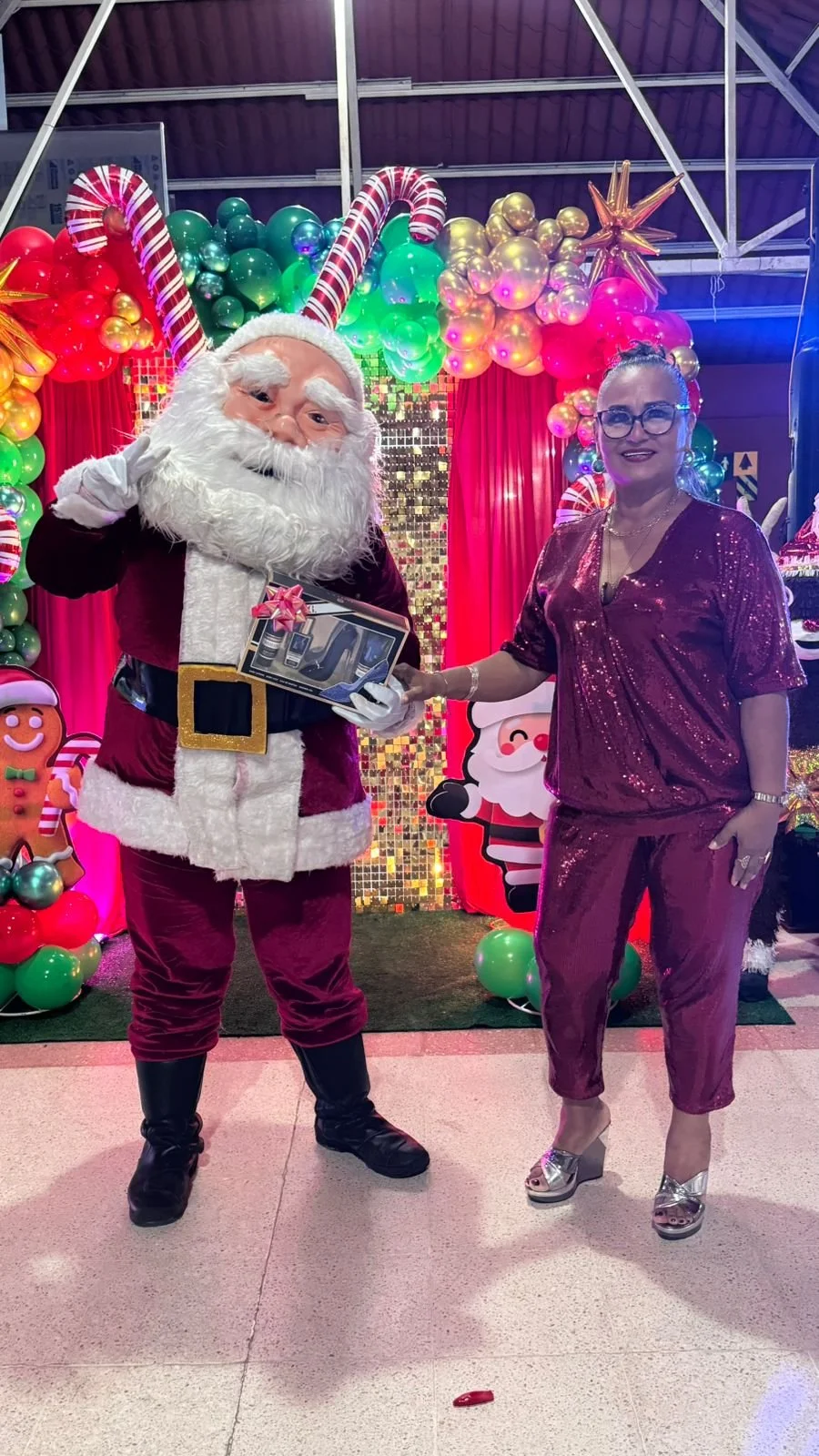 Santa Claus entregando un regalo a una mujer en una decoración navideña con globos, figuras de nieve, Santa, y adornos festivos.