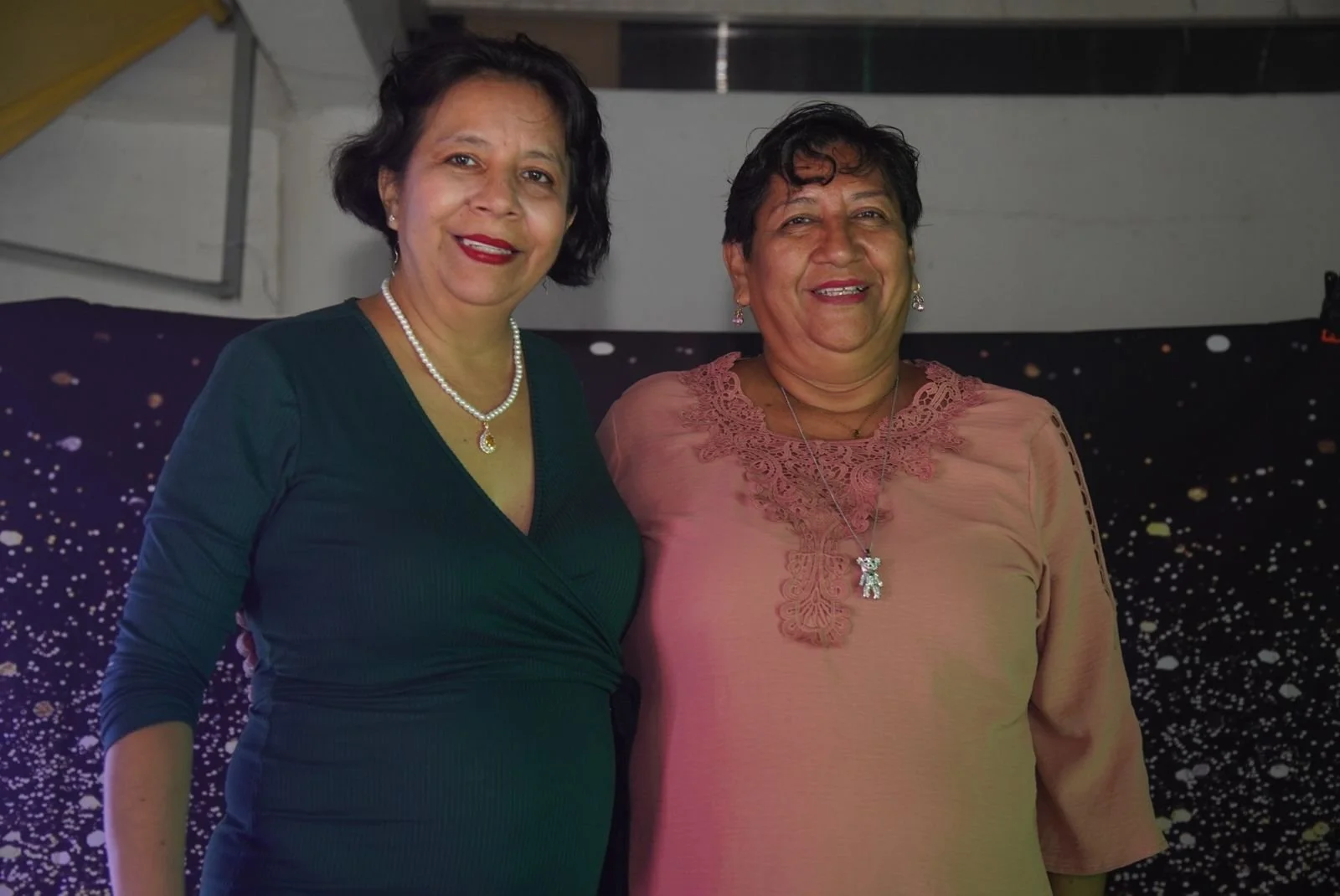 Dos mujeres sonriendo en una celebración, una con un vestido verde y la otra con un blusa rosa claro y accesorios plateados.