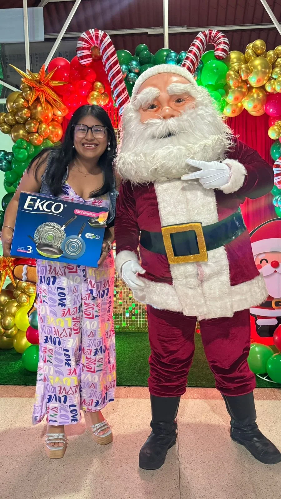 Una mujer con gafas y vestido colorido posando junto a Santa Claus, rodeados de decoraciones navideñas con globos verdes, rojos y dorados, en una escena festiva.