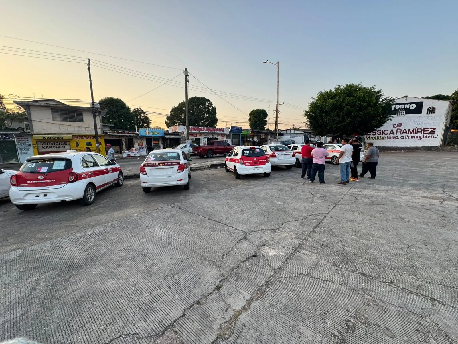 Estacionamiento al aire libre con varios autos blancos y rojos y un grupo de personas conversando. En el fondo, pequeños comercios y un muro con cartel publicitario en un entorno urbano.