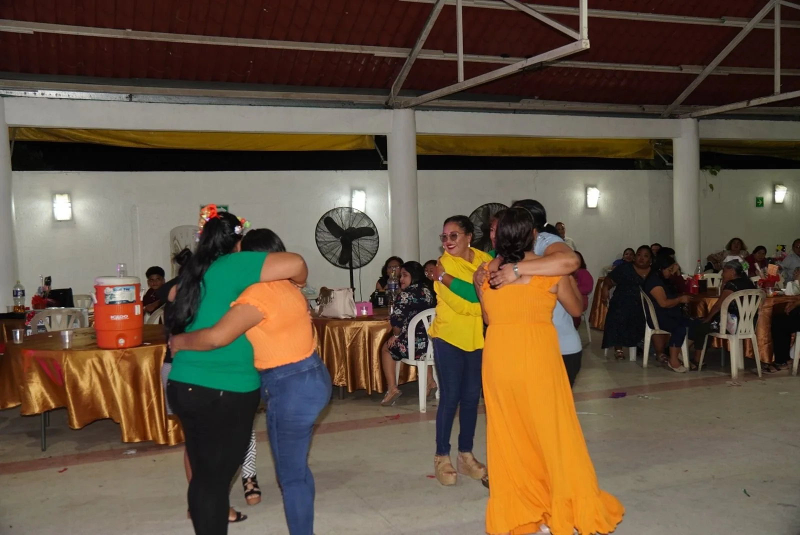 Personas bailando en una fiesta con mesas y asistentes en el fondo.