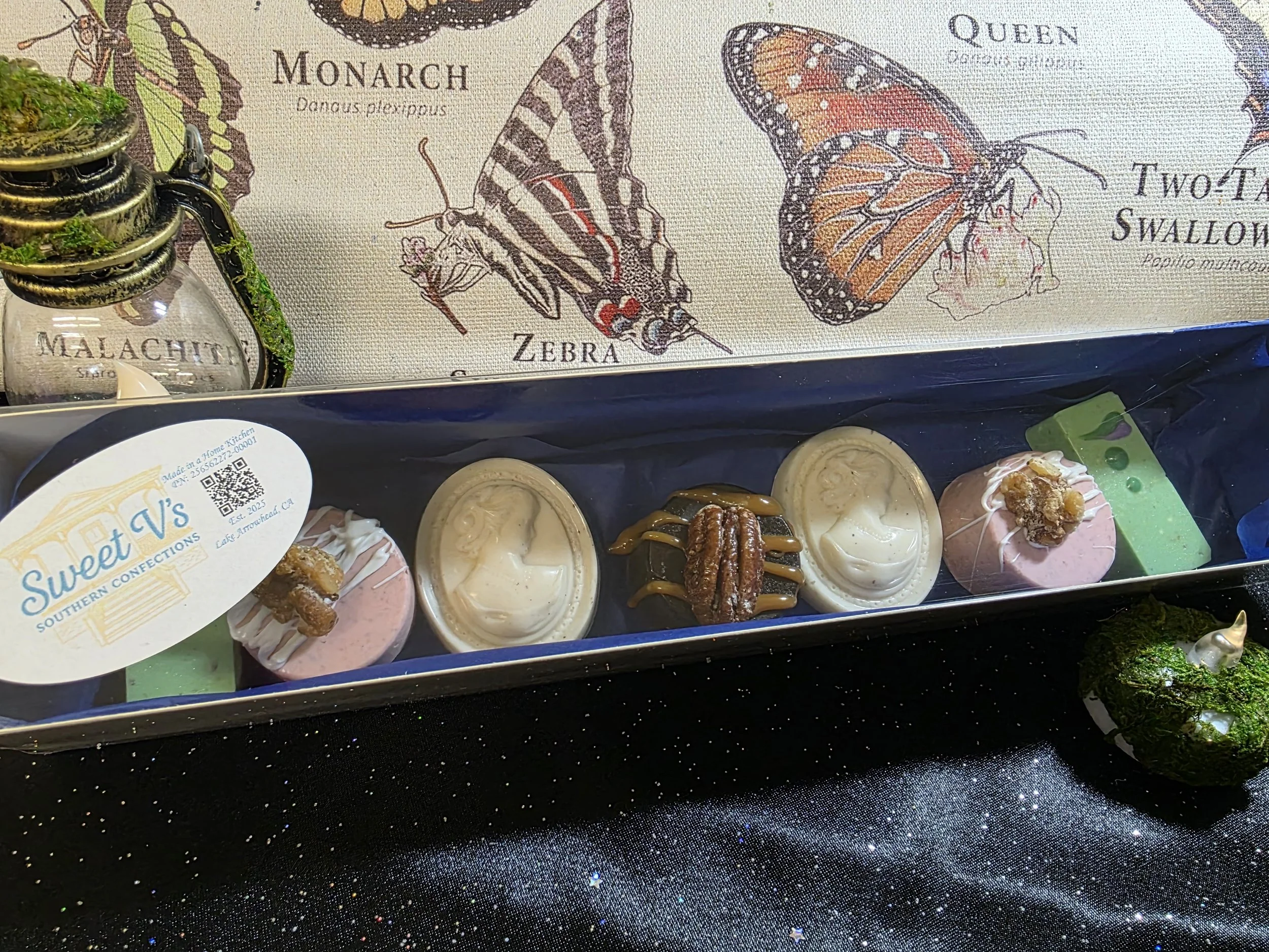 A box of assorted miniature confections with various designs and toppings, placed on a sparkly black surface. Behind the box, there is a decorative illustration of butterflies labeled 'Monarch,' 'Zebra,' 'Queen,' and 'Two Tailed Swallowtail,' with a