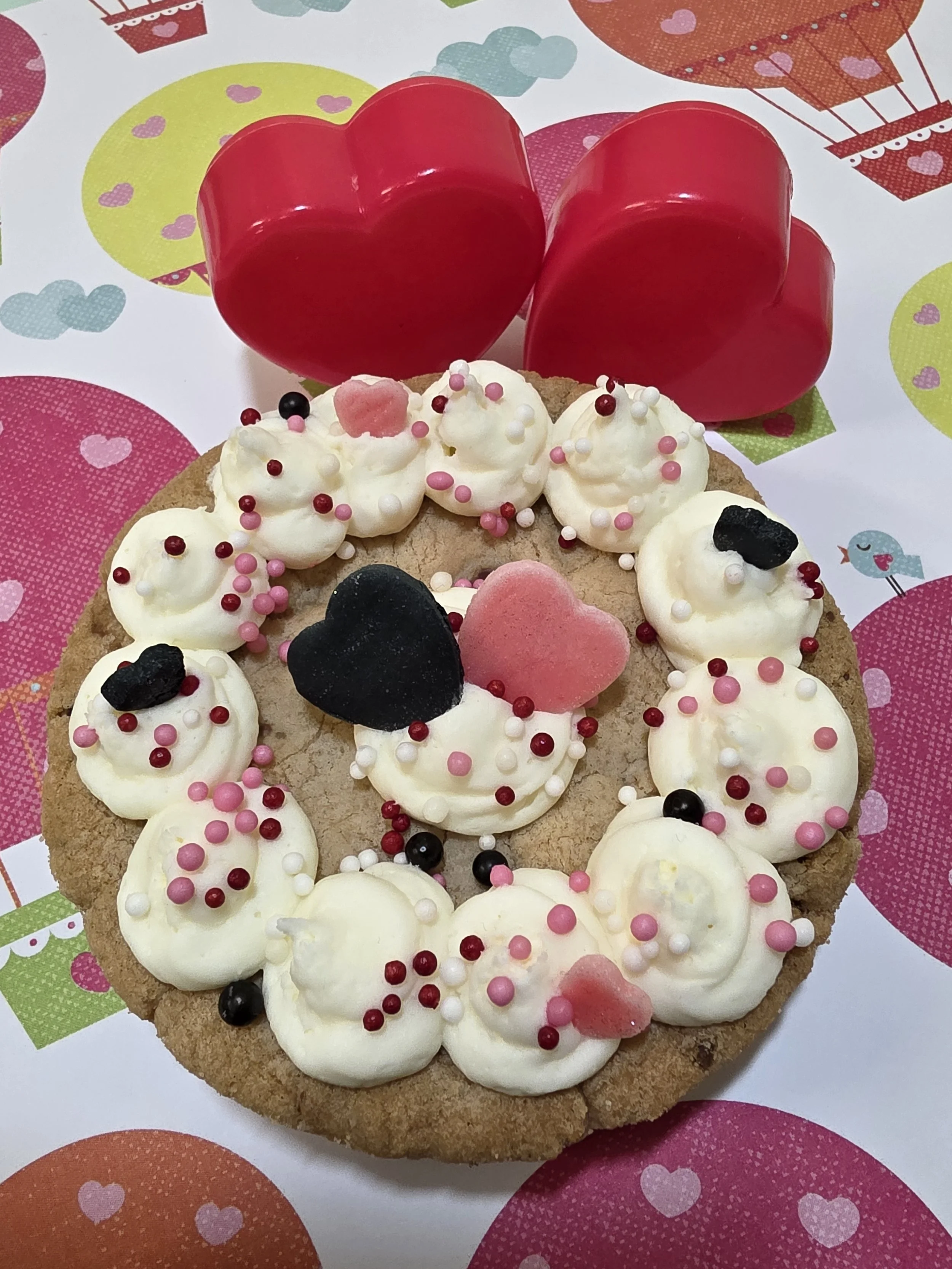 A cookie cake decorated with white frosting, pink, black, and white sprinkles, pink and black gummy candies, and heart-shaped decorations. Behind the cookie are two red, heart-shaped objects and a background with hearts, clouds, and hot air balloons.
