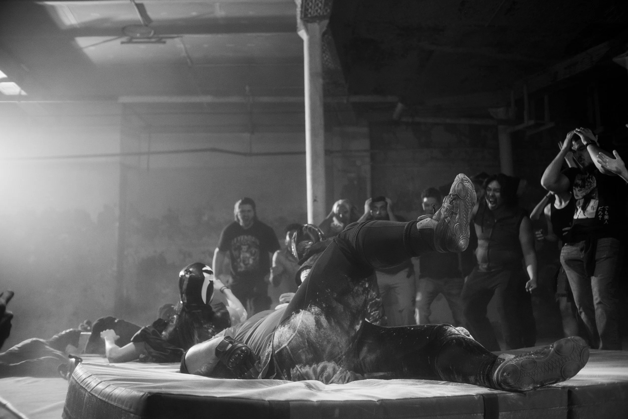 Black and white photo of a wrestling match with masked athletes on padded mat and cheering audience in background.