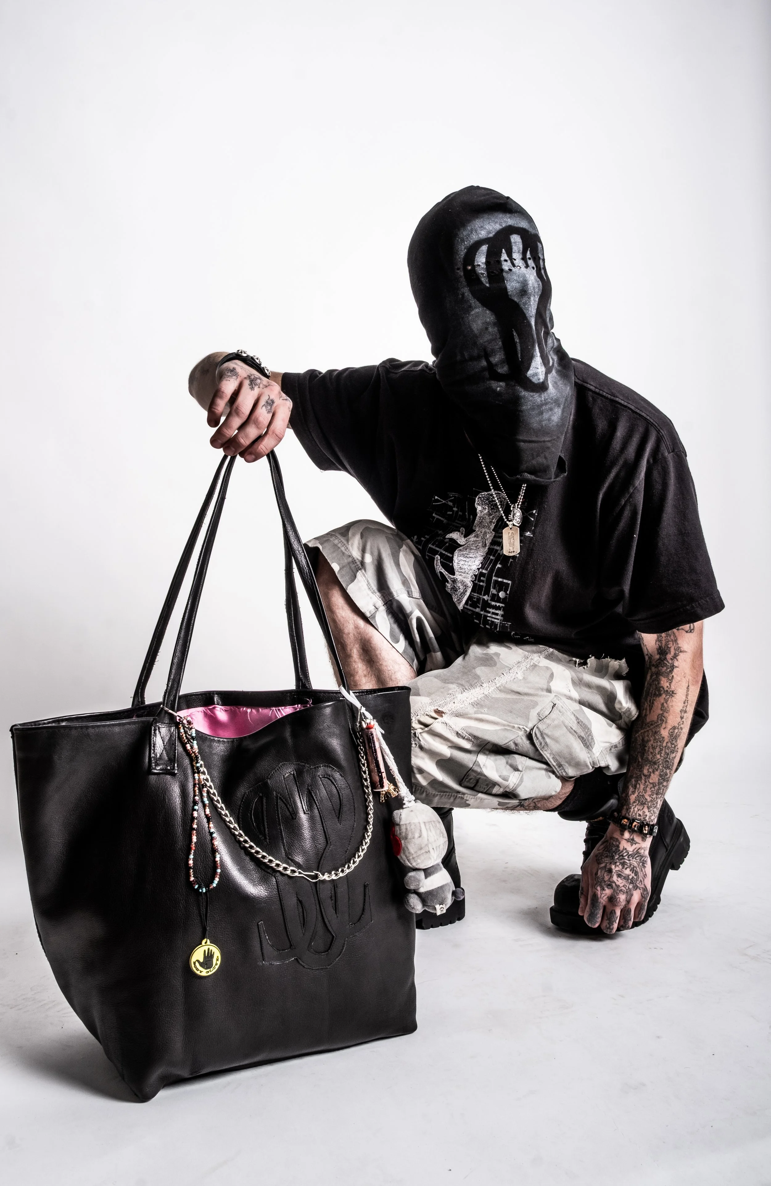 Person wearing a black mask, holding a large black leather bag with decorative chains, kneeling on one knee, dressed in a black shirt and light-colored cargo shorts, tattoos on hands.