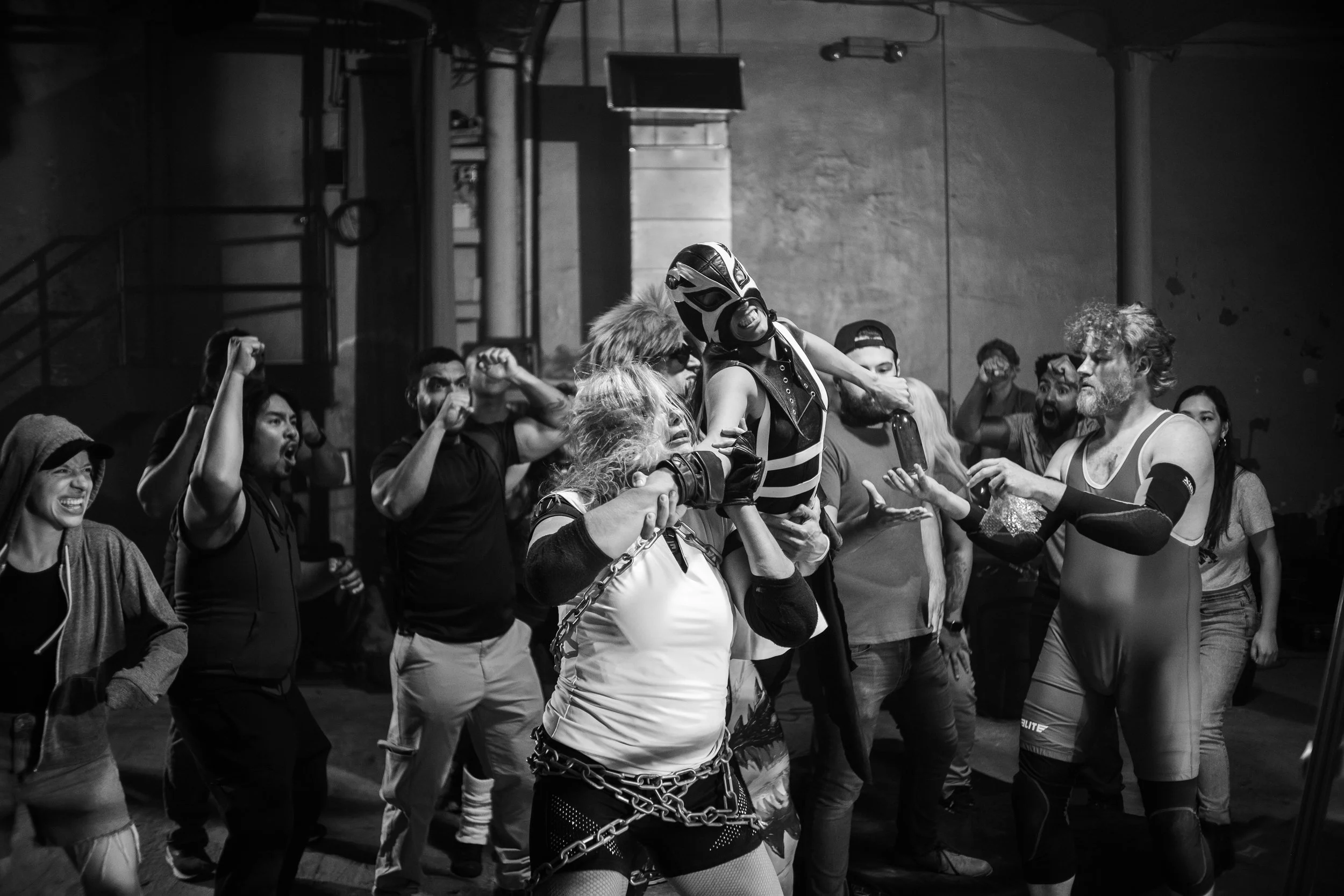 Group of people watching a wrestling match in a dimly lit room, featuring a wrestler lifting another person wearing a mask, surrounded by cheering spectators.