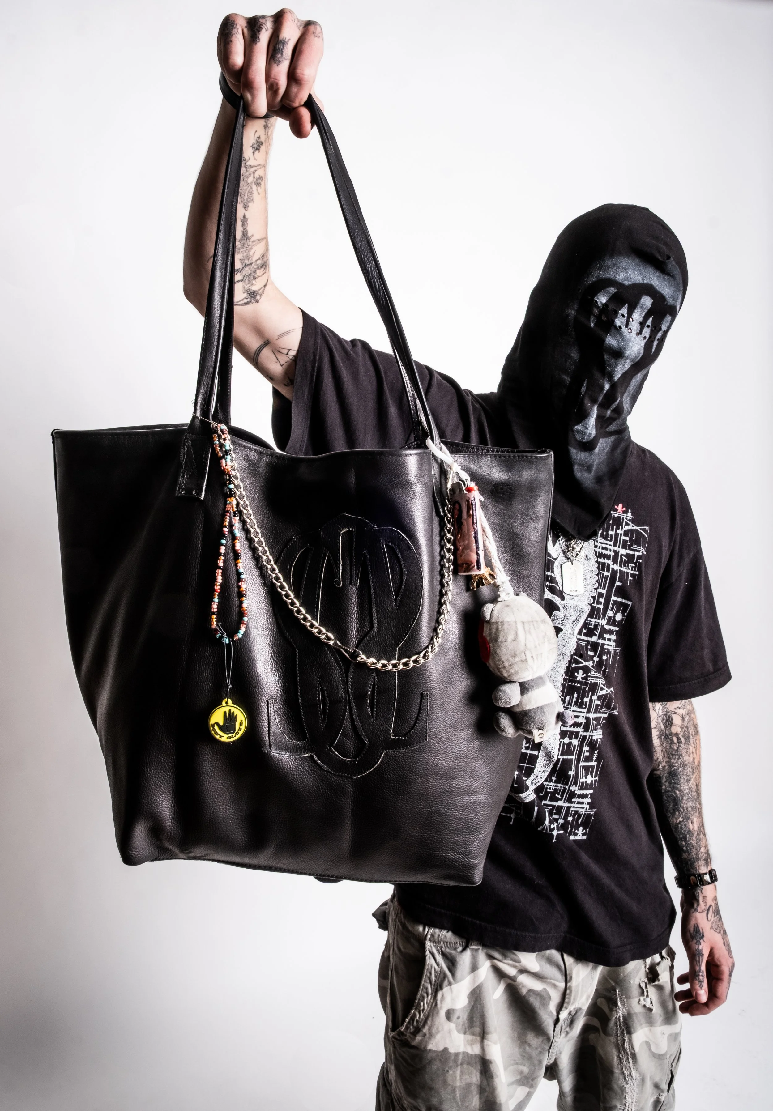 A person wearing a black hood and black shirt holds a large black leather tote bag decorated with charms and a plush toy. The person's arms are tattooed, and they are wearing camouflage pants.