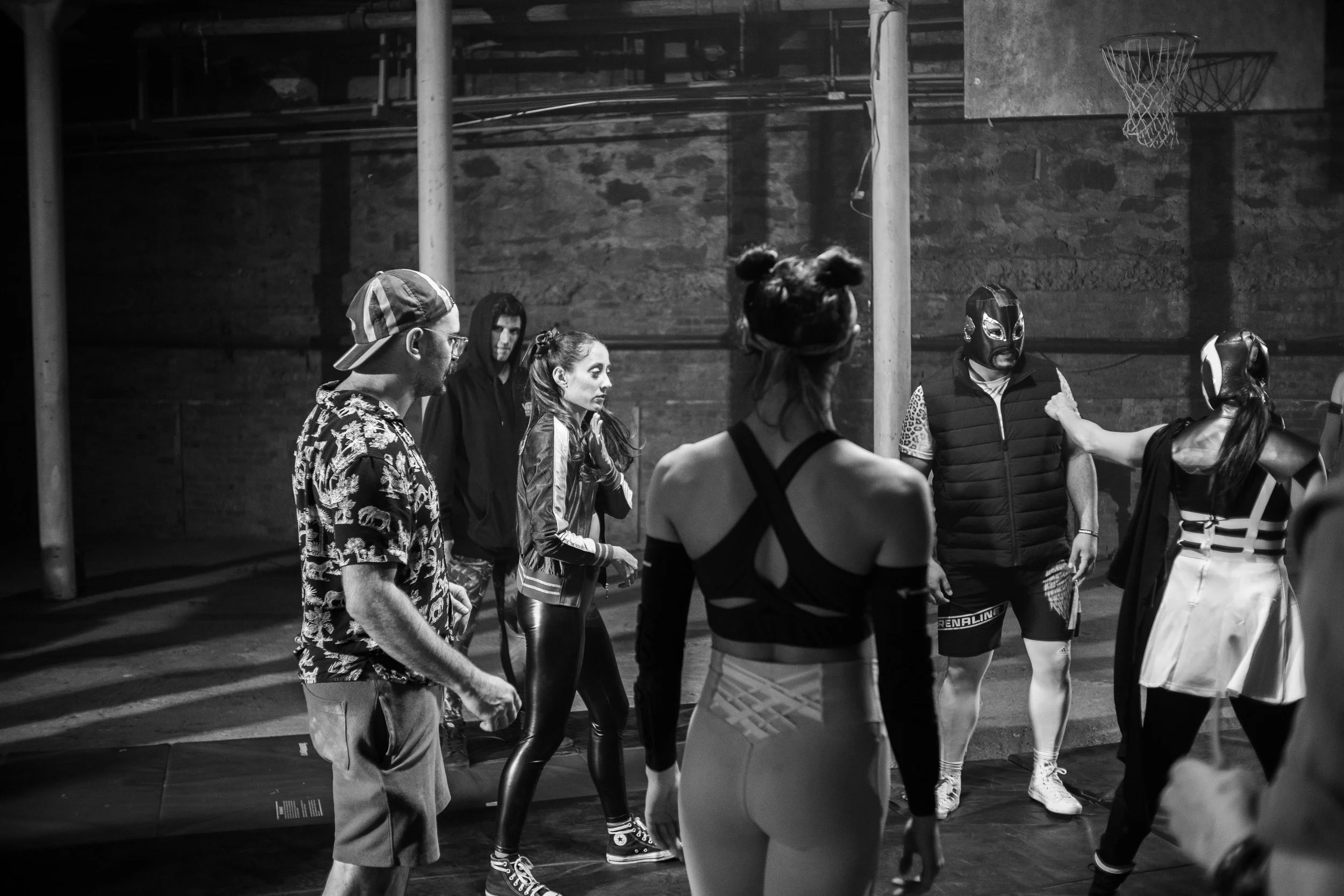 A group of people in a gym setting engaged in an activity. Some are wearing sports attire, including masks and costumes like those worn in wrestling. The setting appears to be a rehearsal or practice, as there is a basketball hoop in the background a