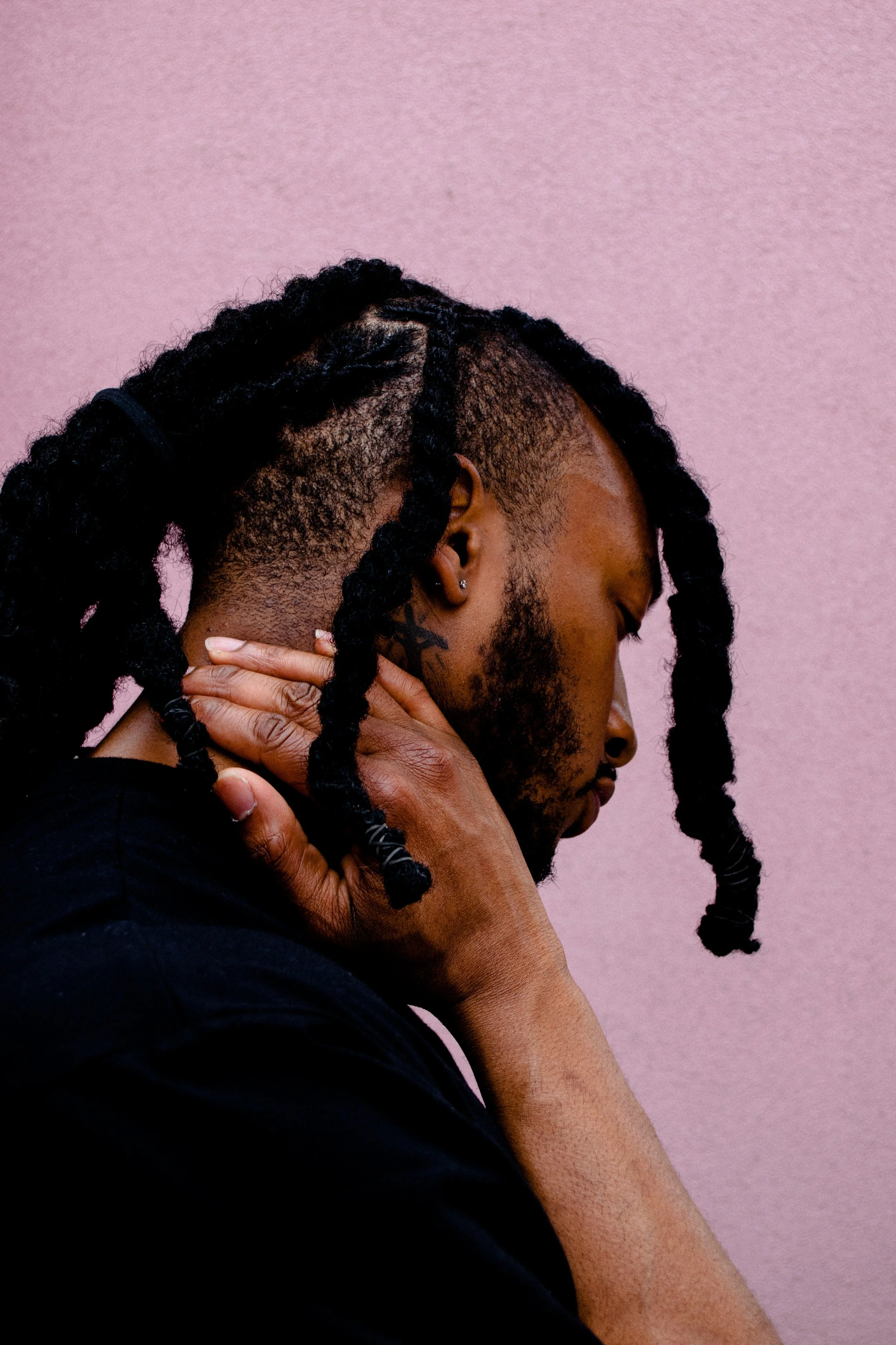 Man with braids holding his neck against a pink background