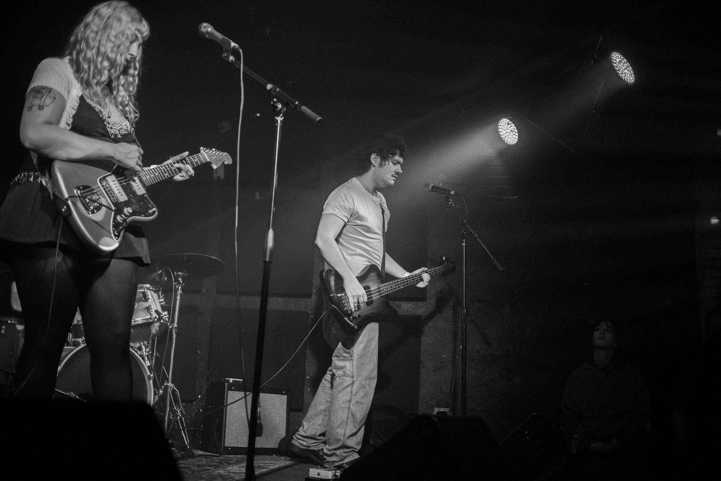 Black and white photo of two musicians on stage, one playing electric guitar and the other playing bass guitar, with microphones and stage lights.