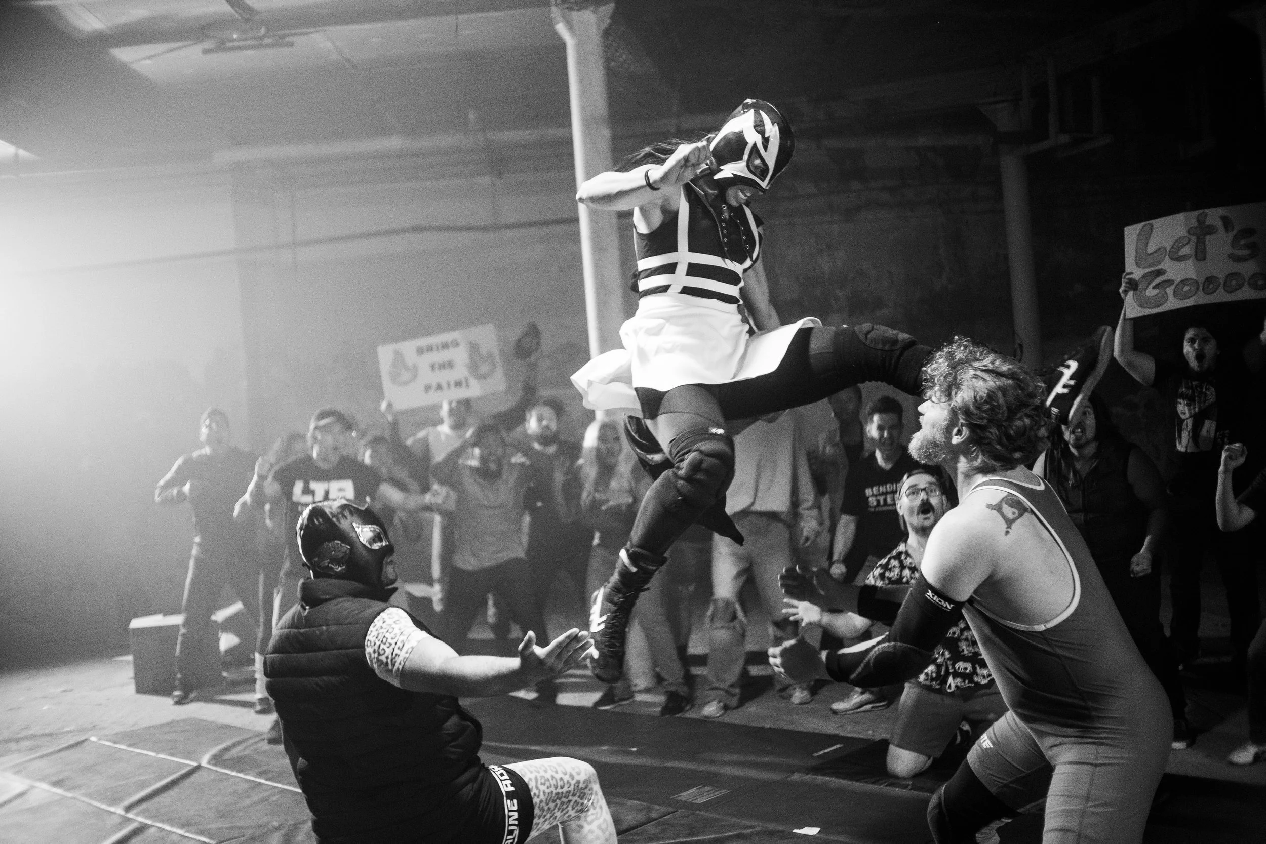 Lucha libre wrestling scene with masked wrestlers performing a move, surrounded by an enthusiastic crowd holding signs, in a dimly lit venue.