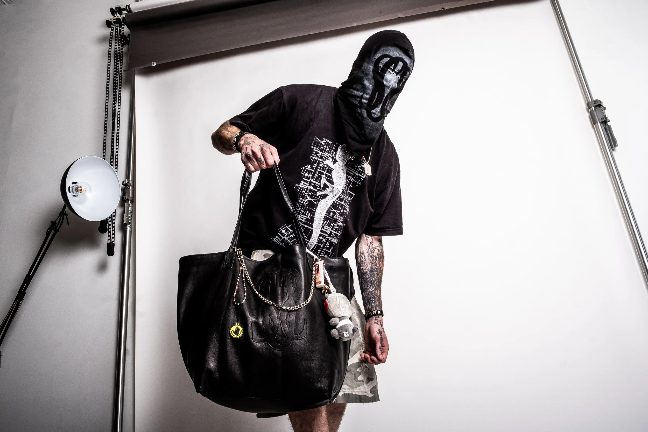Person in black mask holding large black bag with designs, wearing a printed t-shirt, standing in front of a studio backdrop with lighting equipment.