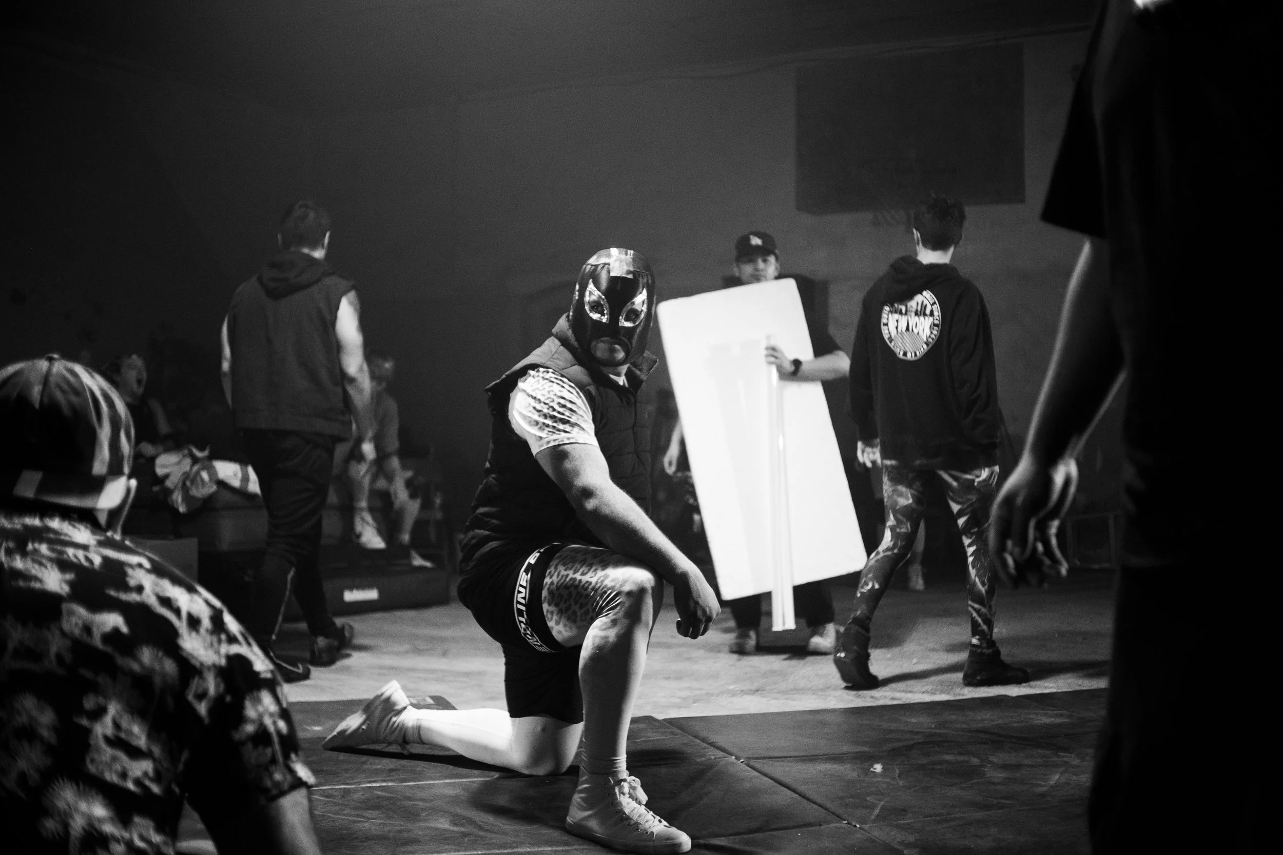 Black and white image of a wrestling scene with a masked wrestler kneeling on the floor, surrounded by other people, in a dimly lit arena.