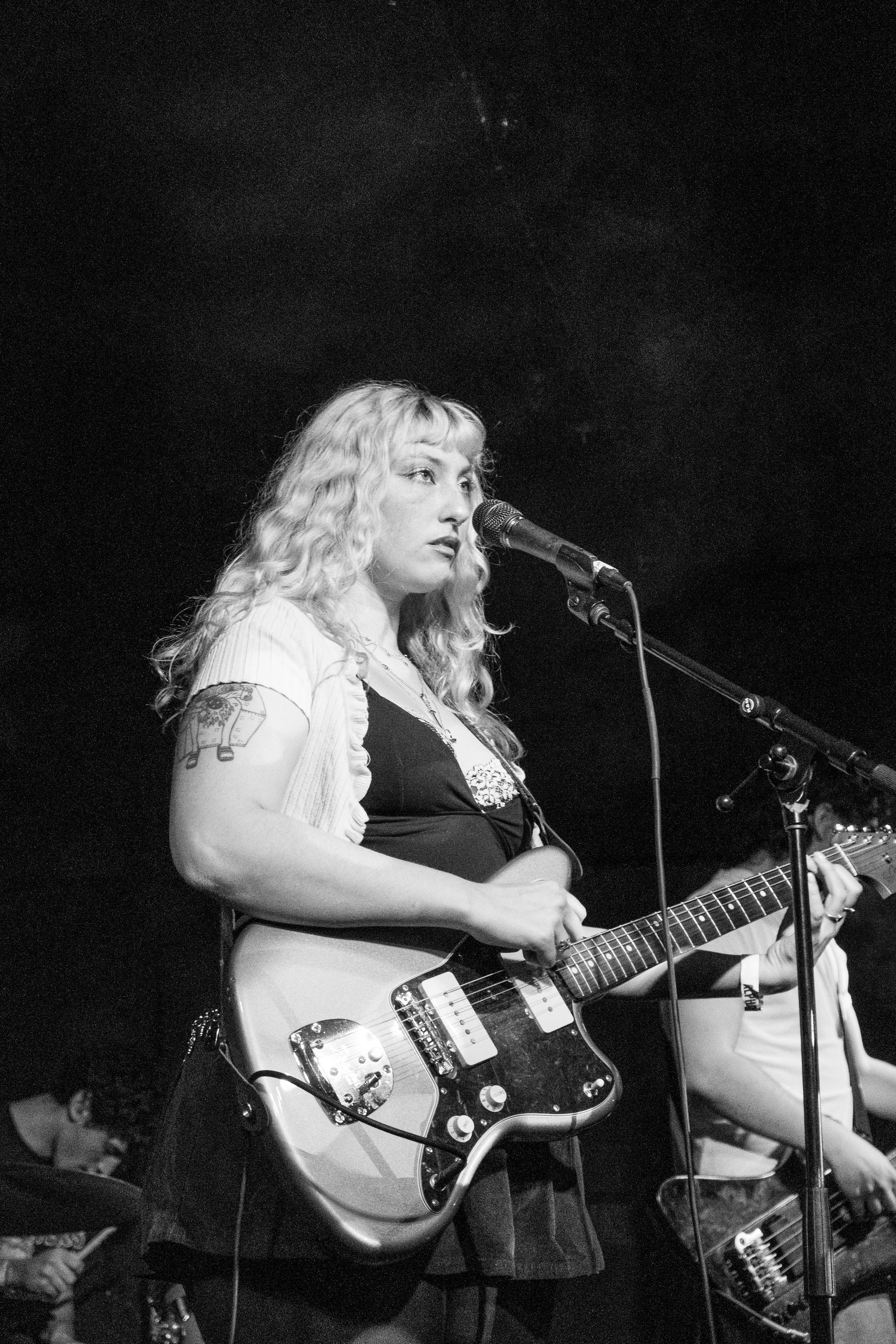 Black and white photo of a woman performing on stage with an electric guitar, singing into a microphone. She has long curly hair and a tattoo on her arm. A guitarist and drummer are in the background.