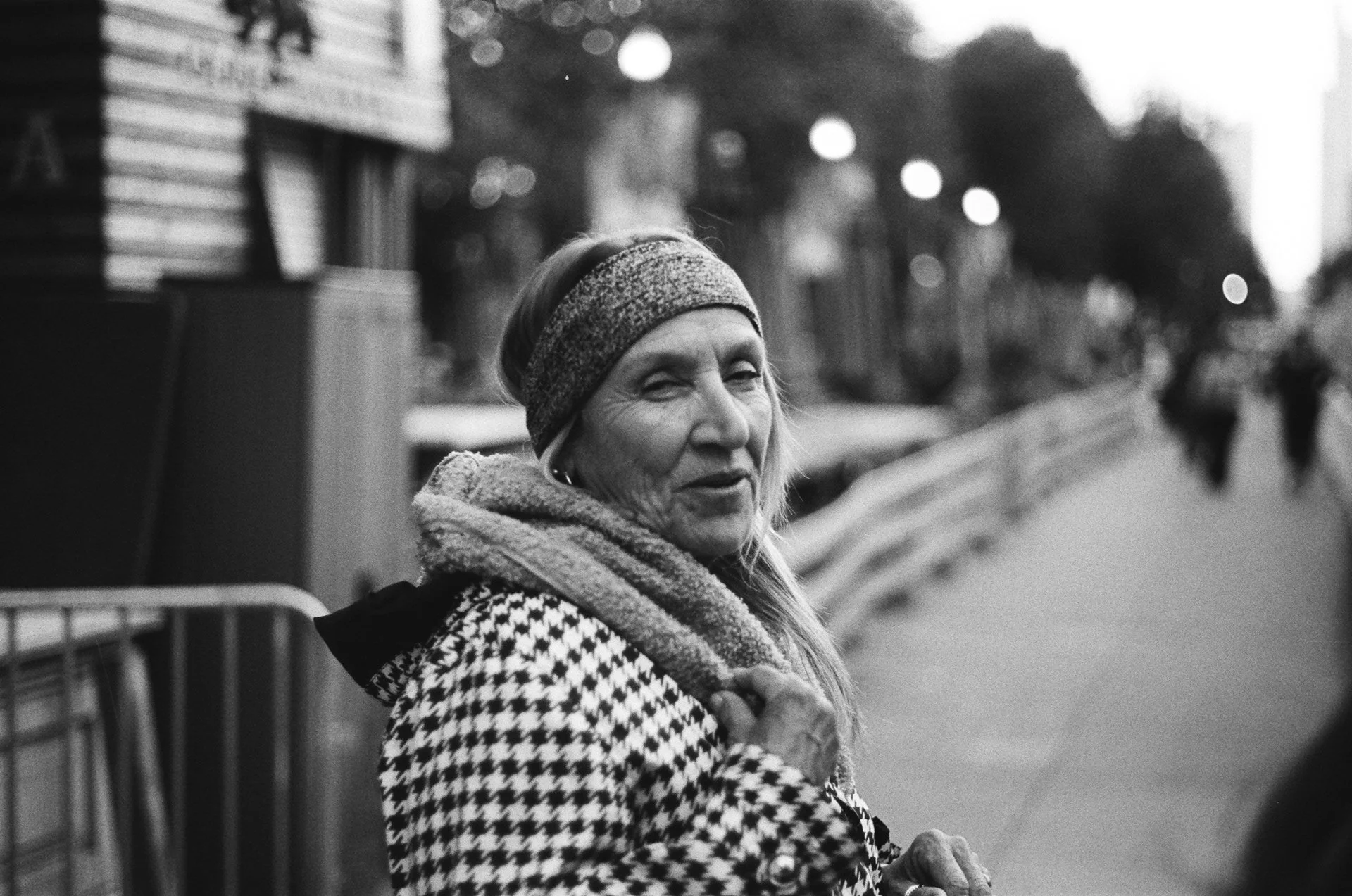 Black and white photo of a woman wearing a houndstooth coat and headband, smiling while walking on a city sidewalk.