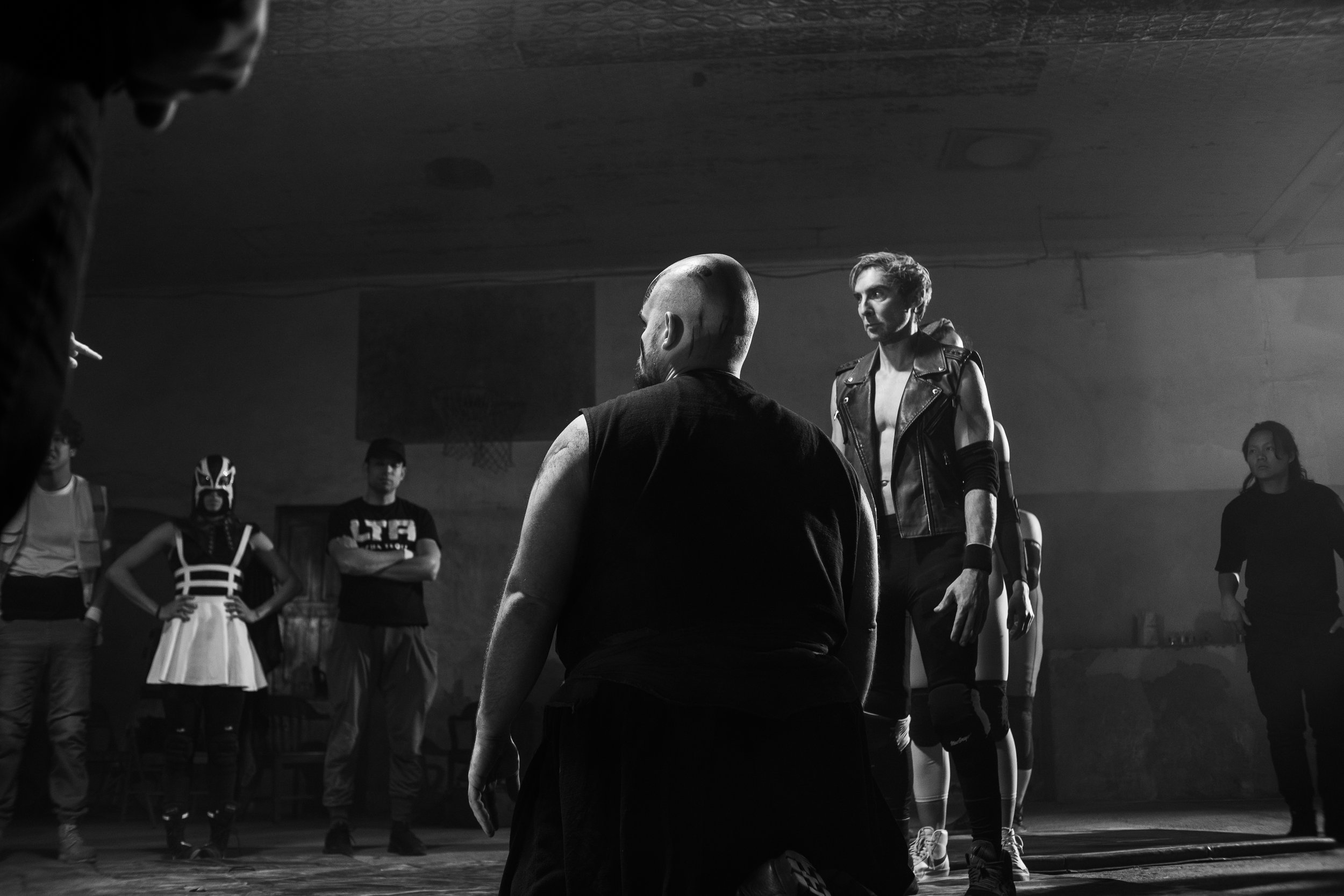 A black-and-white photo shows a tense scene in a dimly lit room with a group of people. One person kneels in the foreground with a mohawk. Others, dressed in eclectic outfits including a wrestling mask and a leather vest, stand watching. The atmosphe