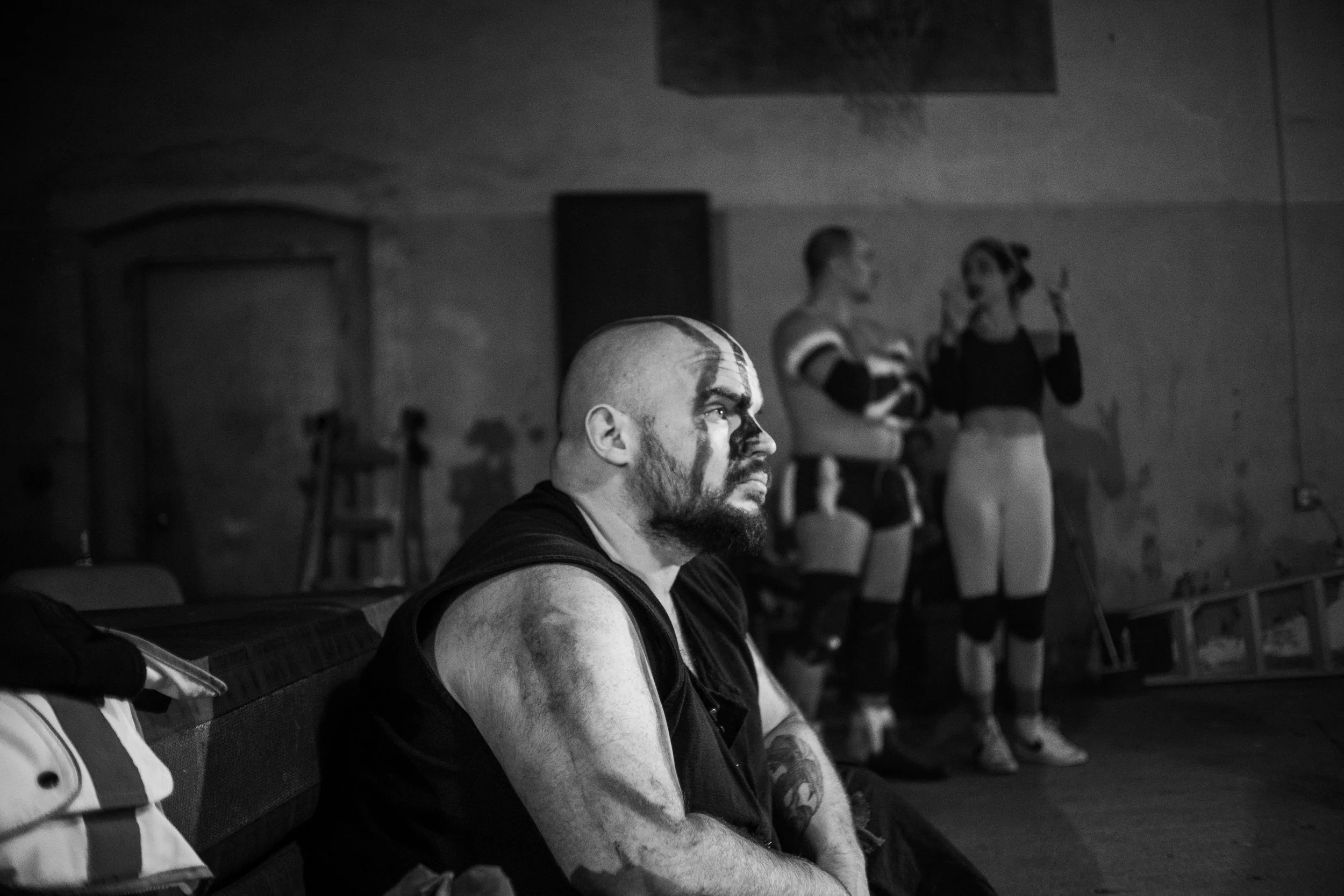 Black and white photo of a man with face paint sitting in a dimly lit room, with two people standing in the background wearing athletic gear. The setting appears to be industrial or gym-like.