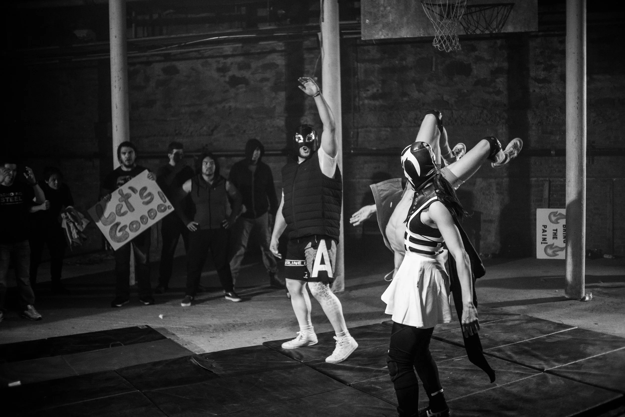 Masked wrestlers performing in a ring with an audience holding signs. One wrestler is lifting another with a wrestling move. "Let's Go" sign visible.