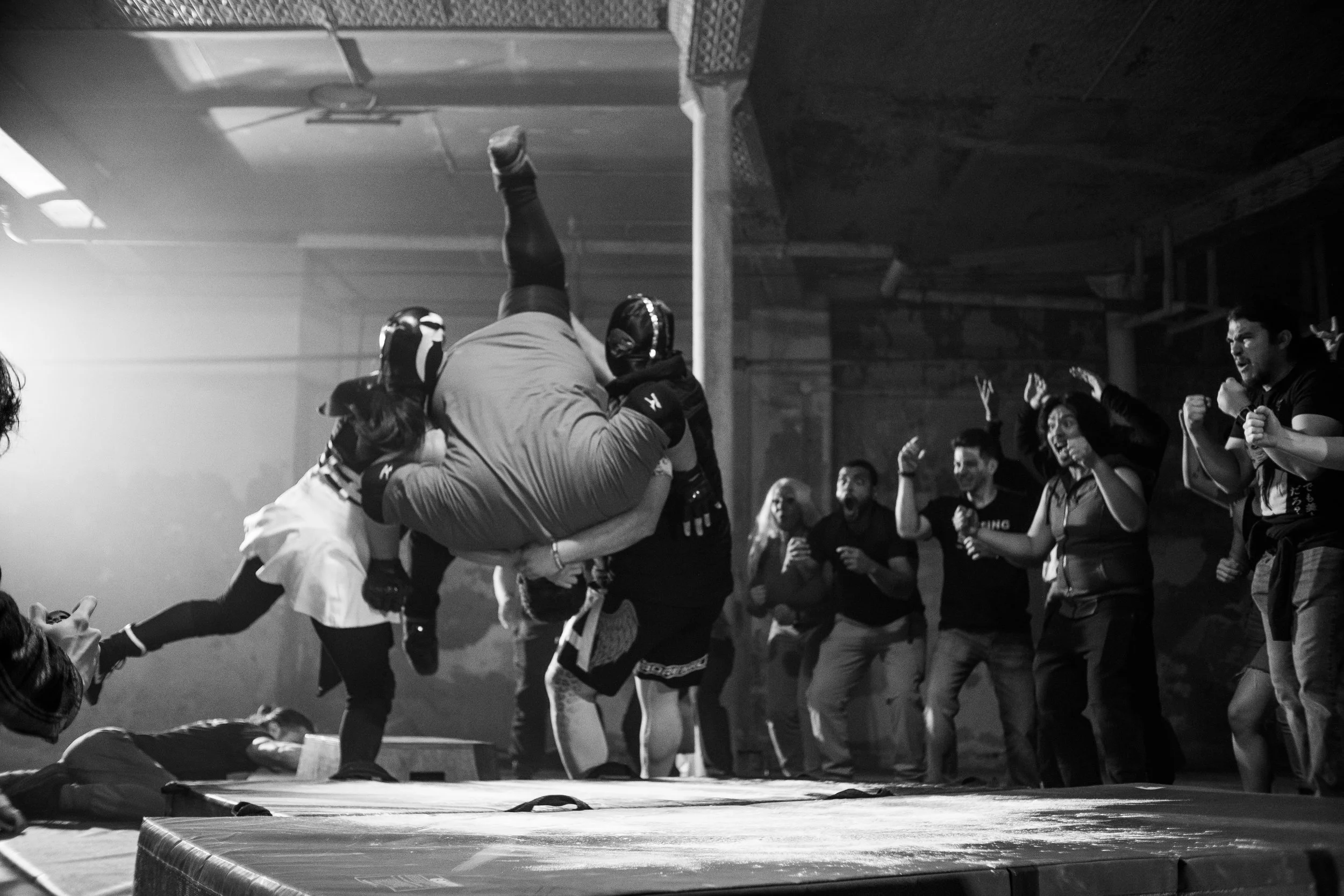 Wrestling match with two masked wrestlers in action, one lifting the other, surrounded by an excited audience in a dimly lit venue.