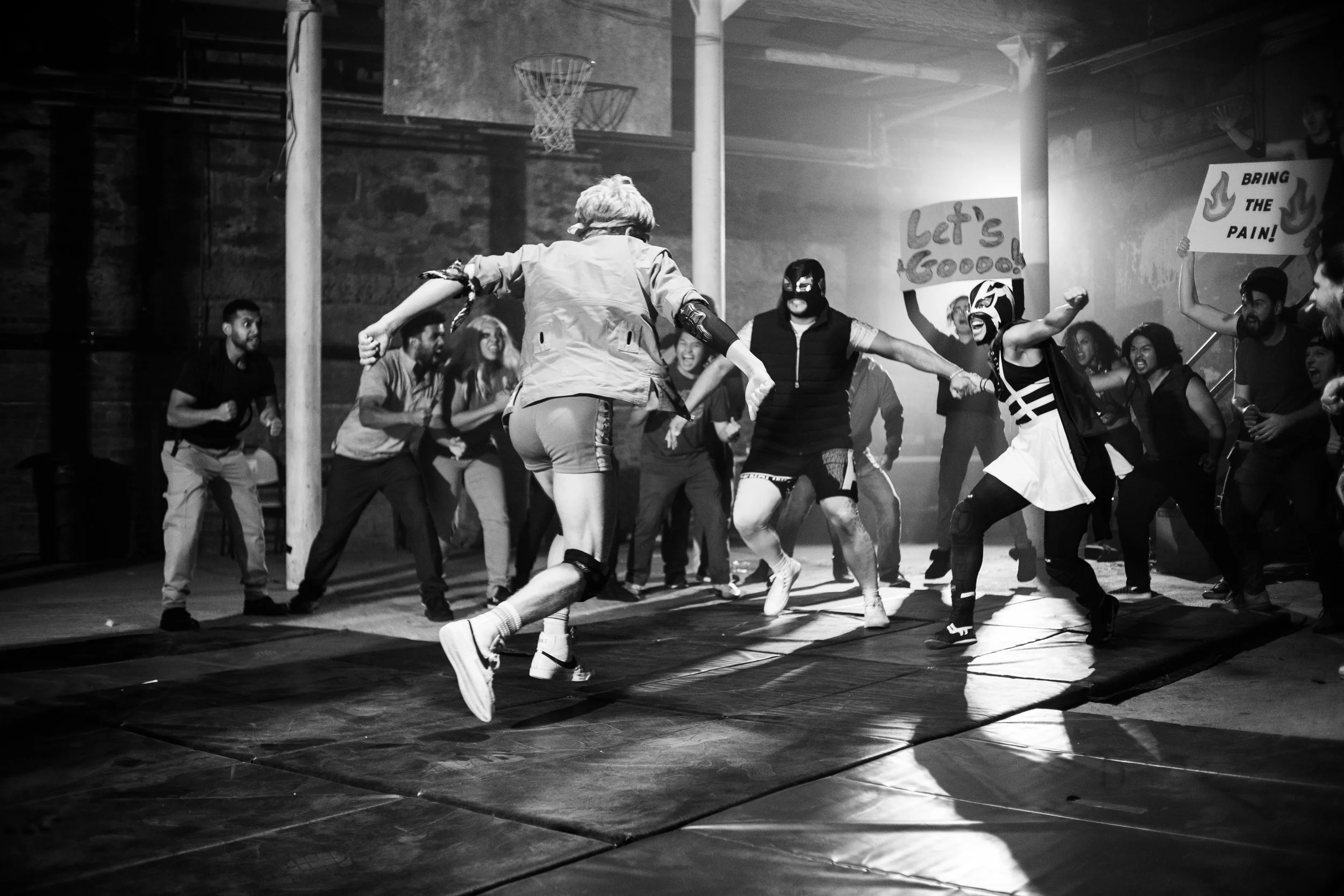 Black and white photo of an indoor wrestling match with costumed participants and an enthusiastic crowd holding signs.