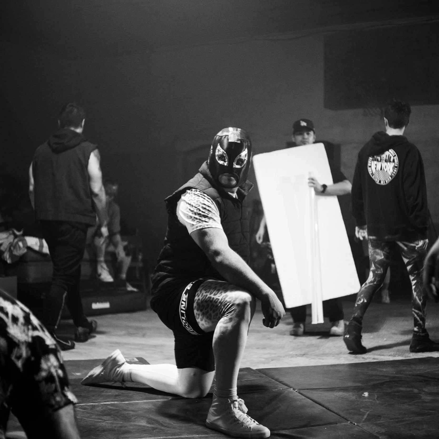 Black and white image of a masked wrestler kneeling in a wrestling ring with several people standing in the background.