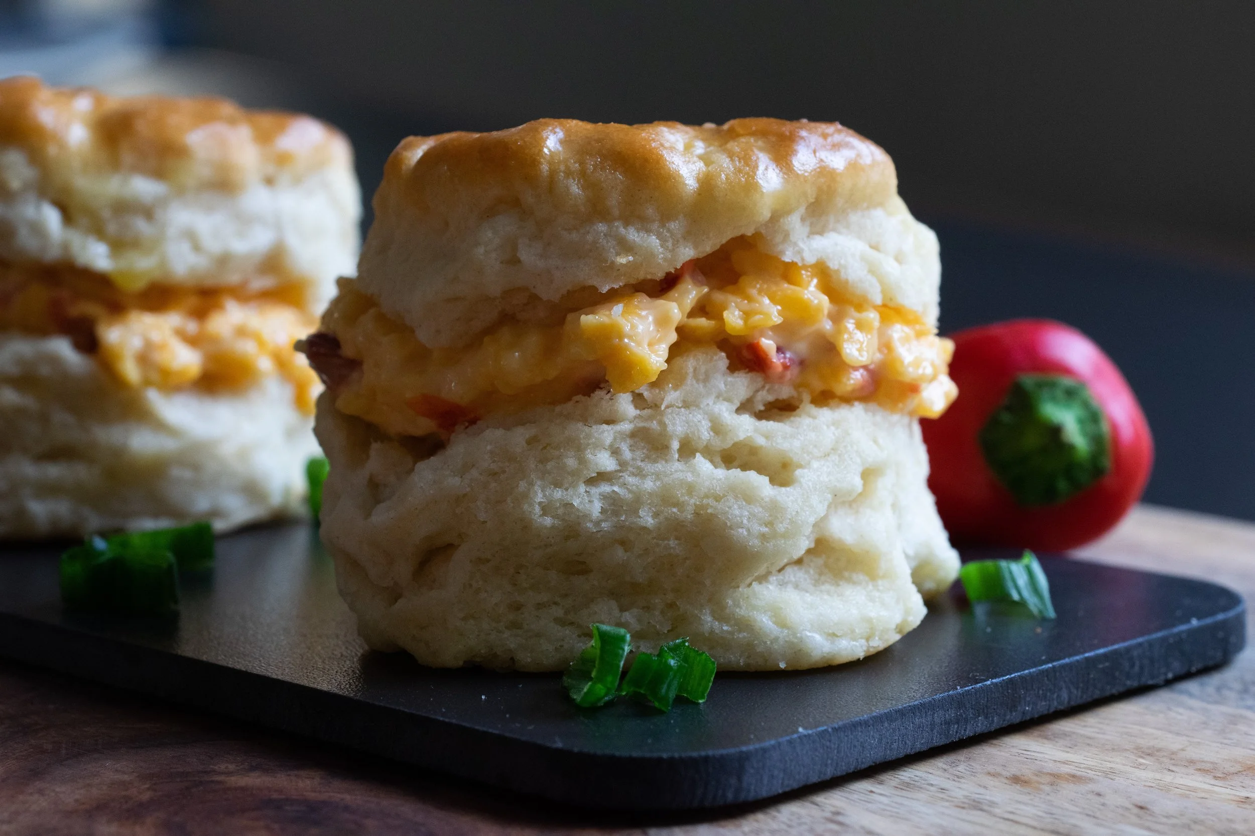 Close-up of a biscuit sandwich filled with pimento cheese, placed on a black surface with chopped green onions and a red chili pepper in the background.