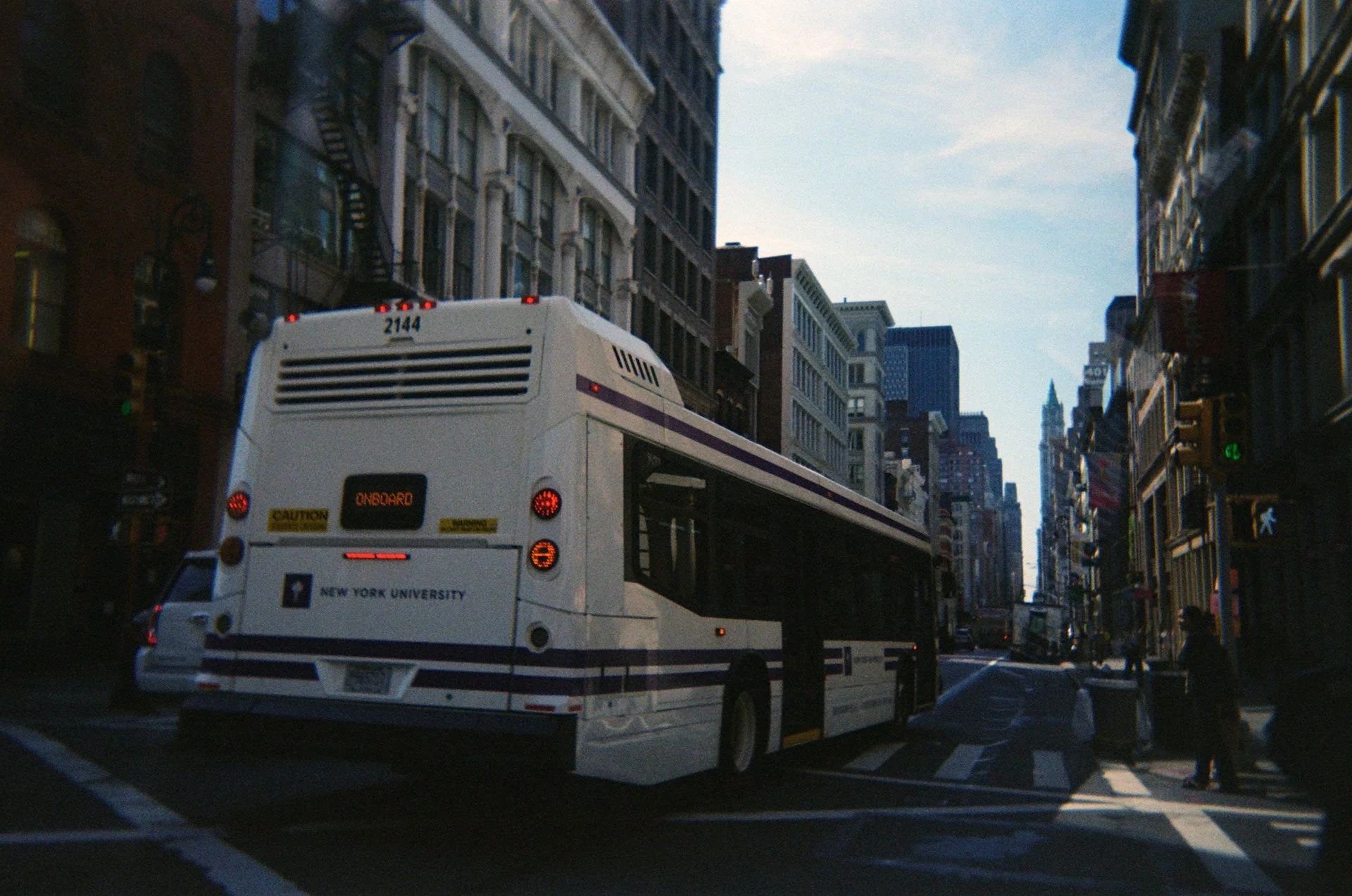 City street with a New York University shuttle bus, surrounded by tall buildings and pedestrians.