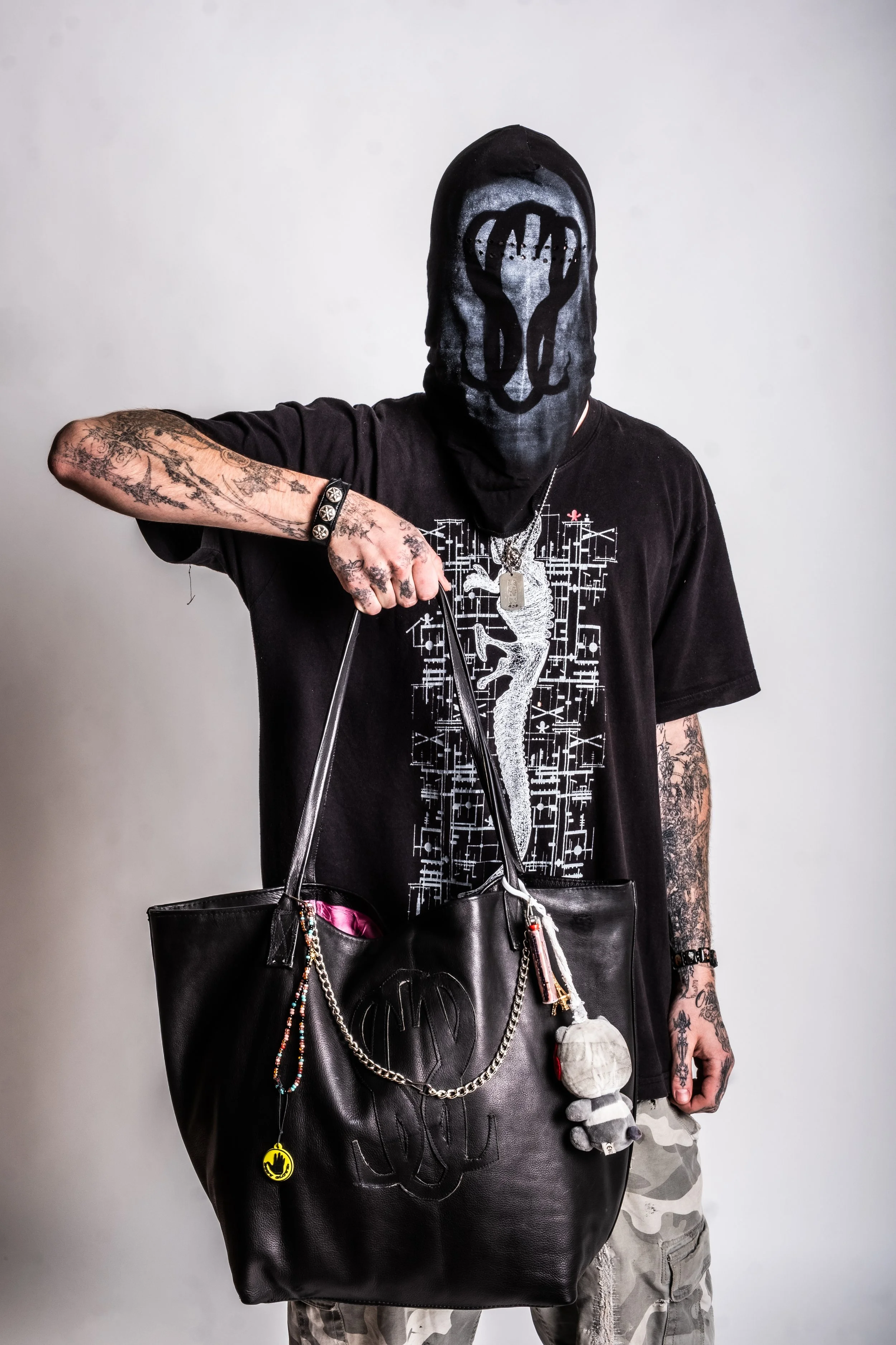 Person with tattoos wearing a mask and t-shirt, holding a black leather tote bag with decorations.
